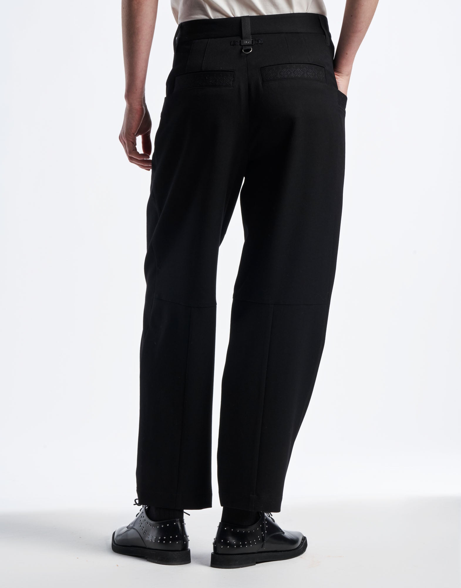 Rear view of a person wearing the black COMPLY pants. The pants feature double darts at the waist and two welt pockets embroidered with a geometric pattern.