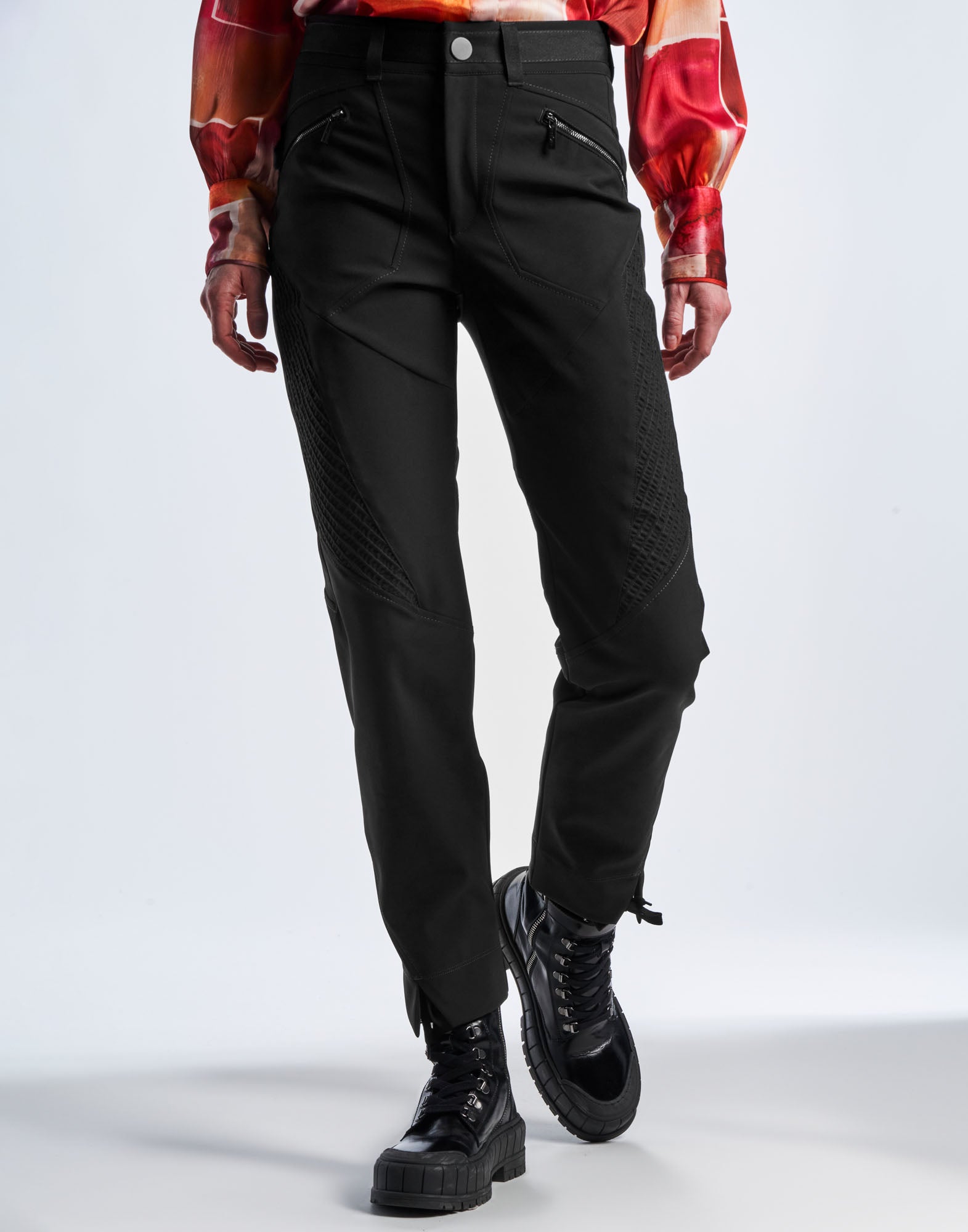 Front view of DEVIATE pants in black tech twill, showing visible contrasting topstitching, diagonal motocross-inspired details, and front zip pockets.