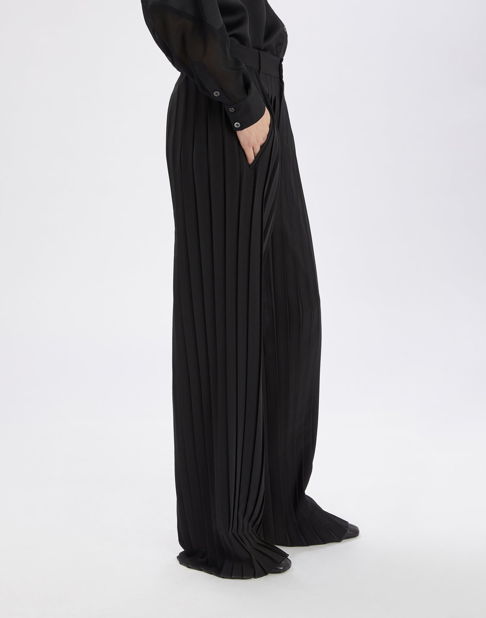 Side view of DREW palazzo trousers in black with deep pleats, straight leg, belt loops, and diagonal front pockets, worn by a person.
