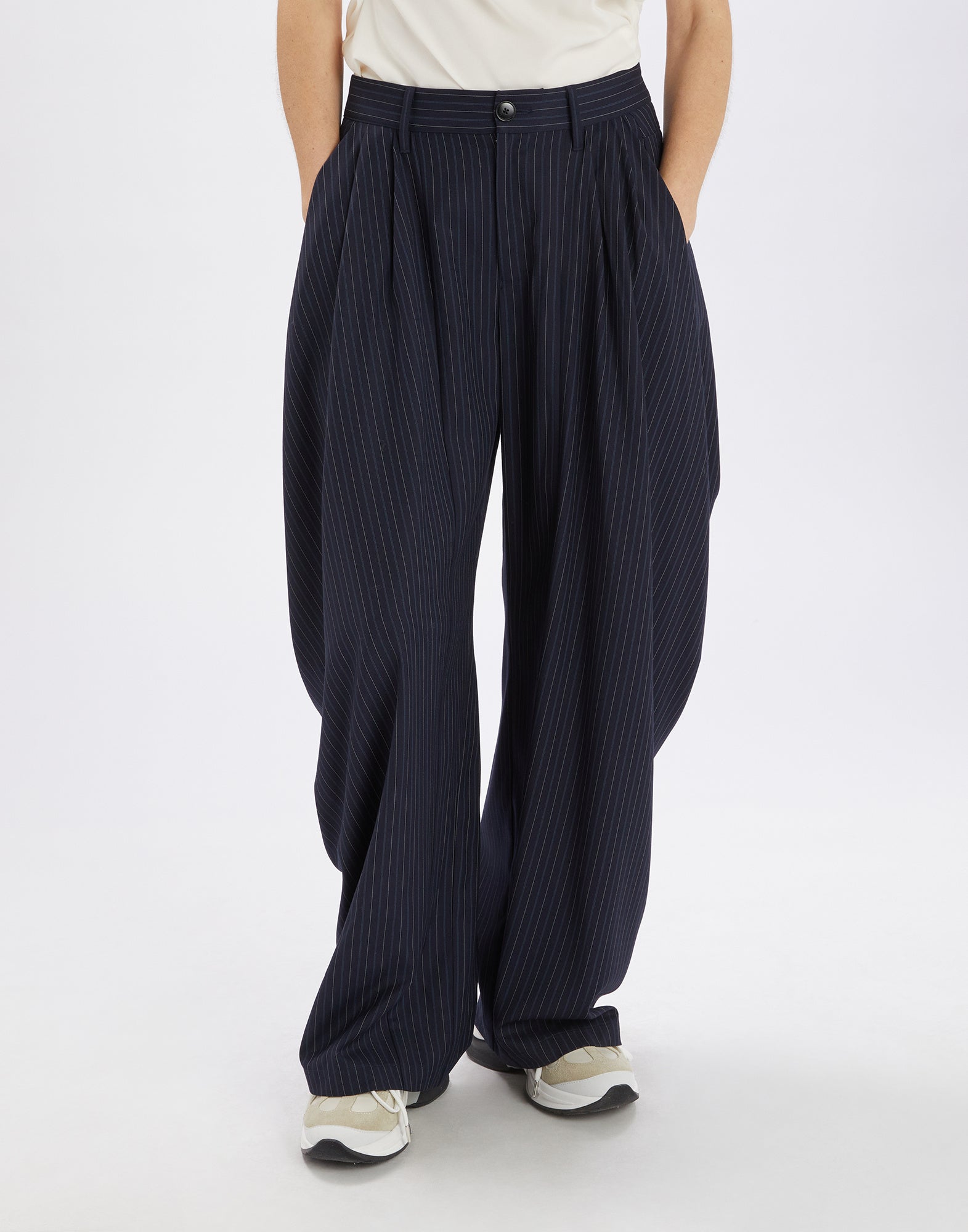 Front view of a person wearing the AMIDST pants; the pants feature a striking drape effect at the hips, fine pinstripes, and a wide cut, combined with a belted waistband and side pockets.