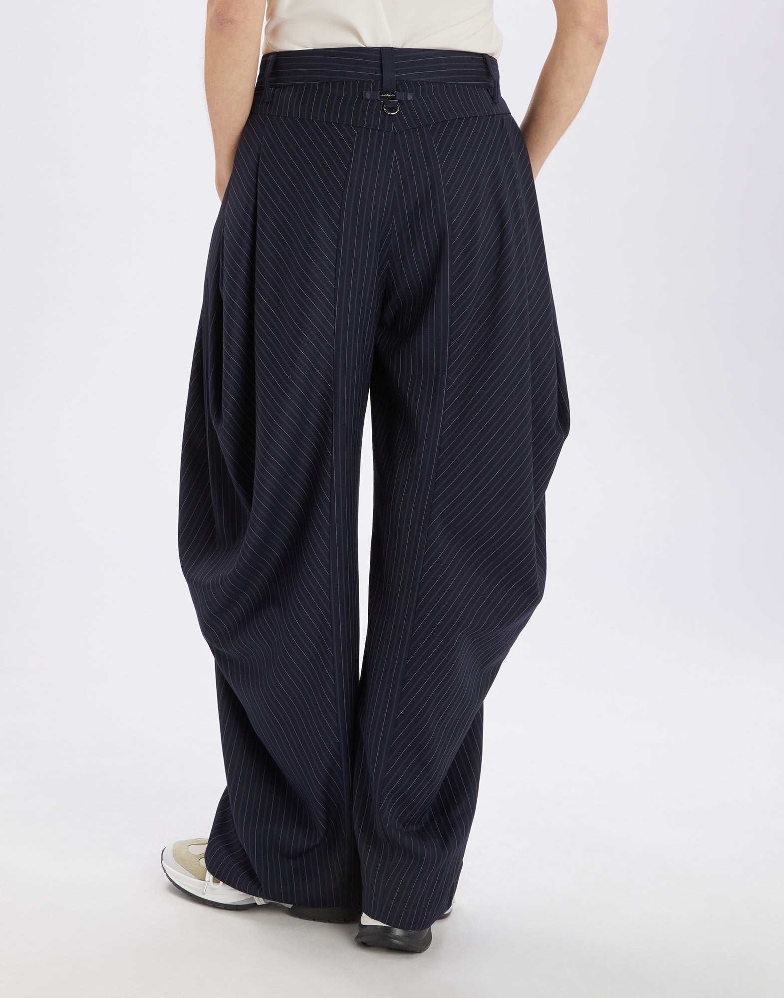 Rear view of AMIDST pants with striking draping at the hips, deep pleats, and a narrow pinstripe pattern in dark blue, worn by a person.
