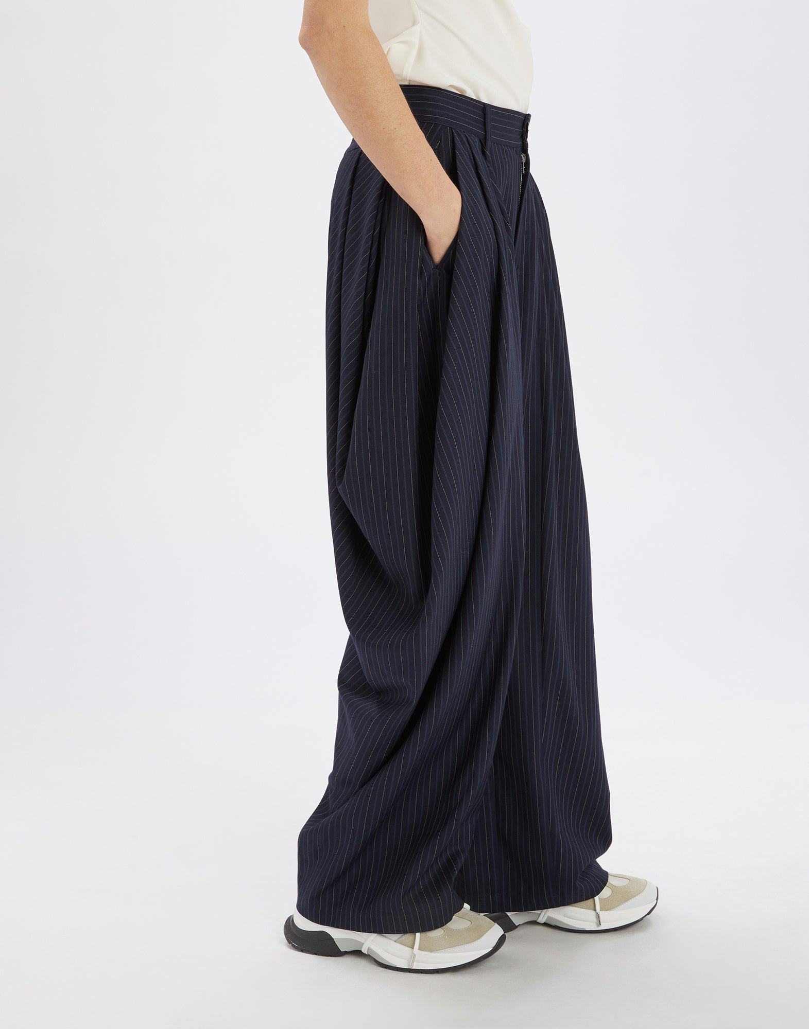 Side view of the AMIDST pants, worn by a person. The pants feature bold draping and deep pleats, made from dark blue striped fabric.