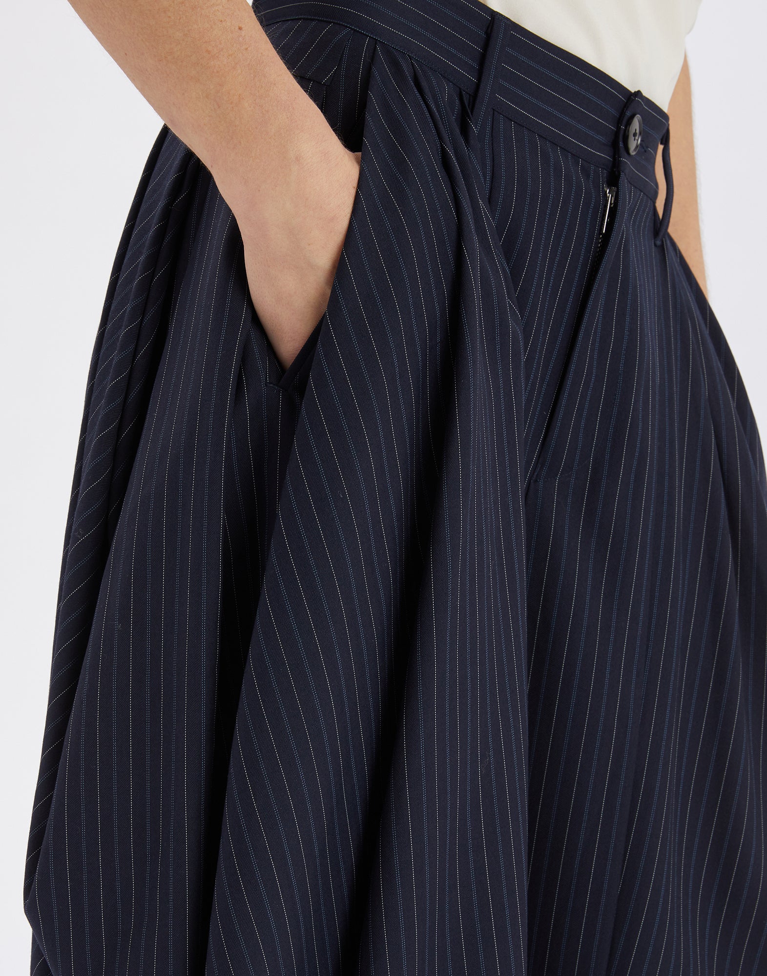 Close-up view of the AMIDST, a classic dark blue office pant with fine pinstripes, bold drape and pleats at the hips, hand in side pocket.