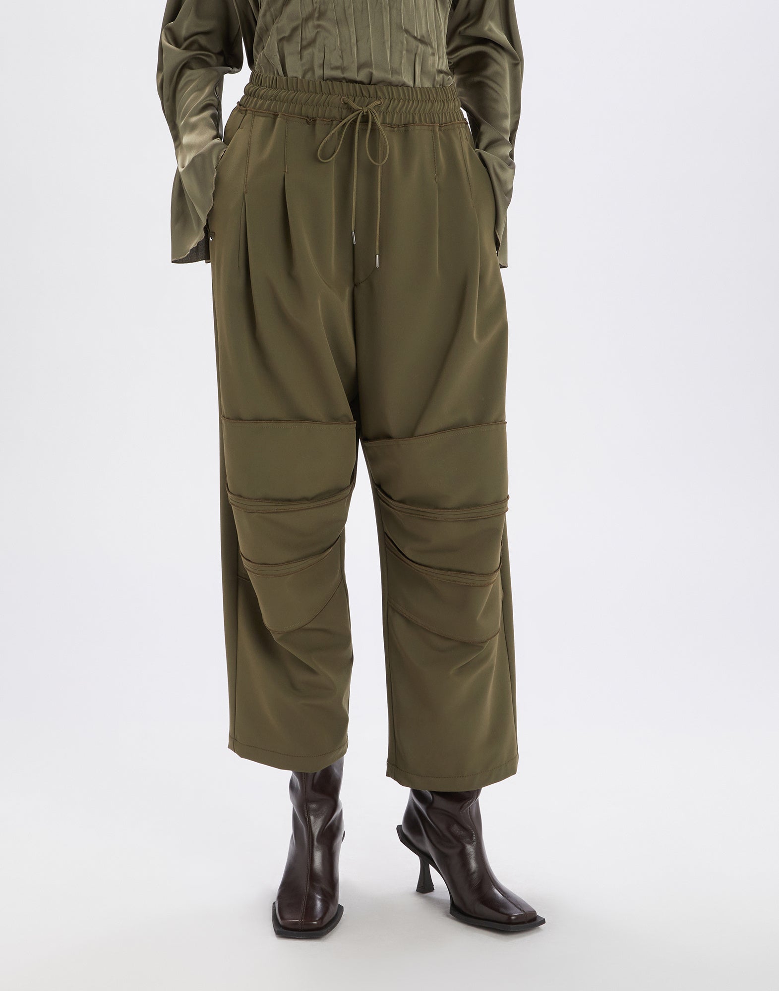 Front view of SHEILA pants in olive green with elastic waistband, drawstring, knee-panel effect, and decorative topstitching, worn with dark ankle boots.