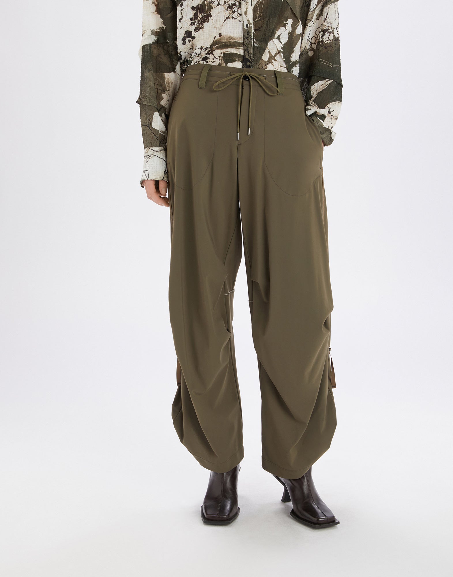 Front view: OUTGOING trousers in olive green, featuring wide-leg cut, drawstring waist, large side pockets, and D-ring detail on the leg, paired with dark ankle boots.