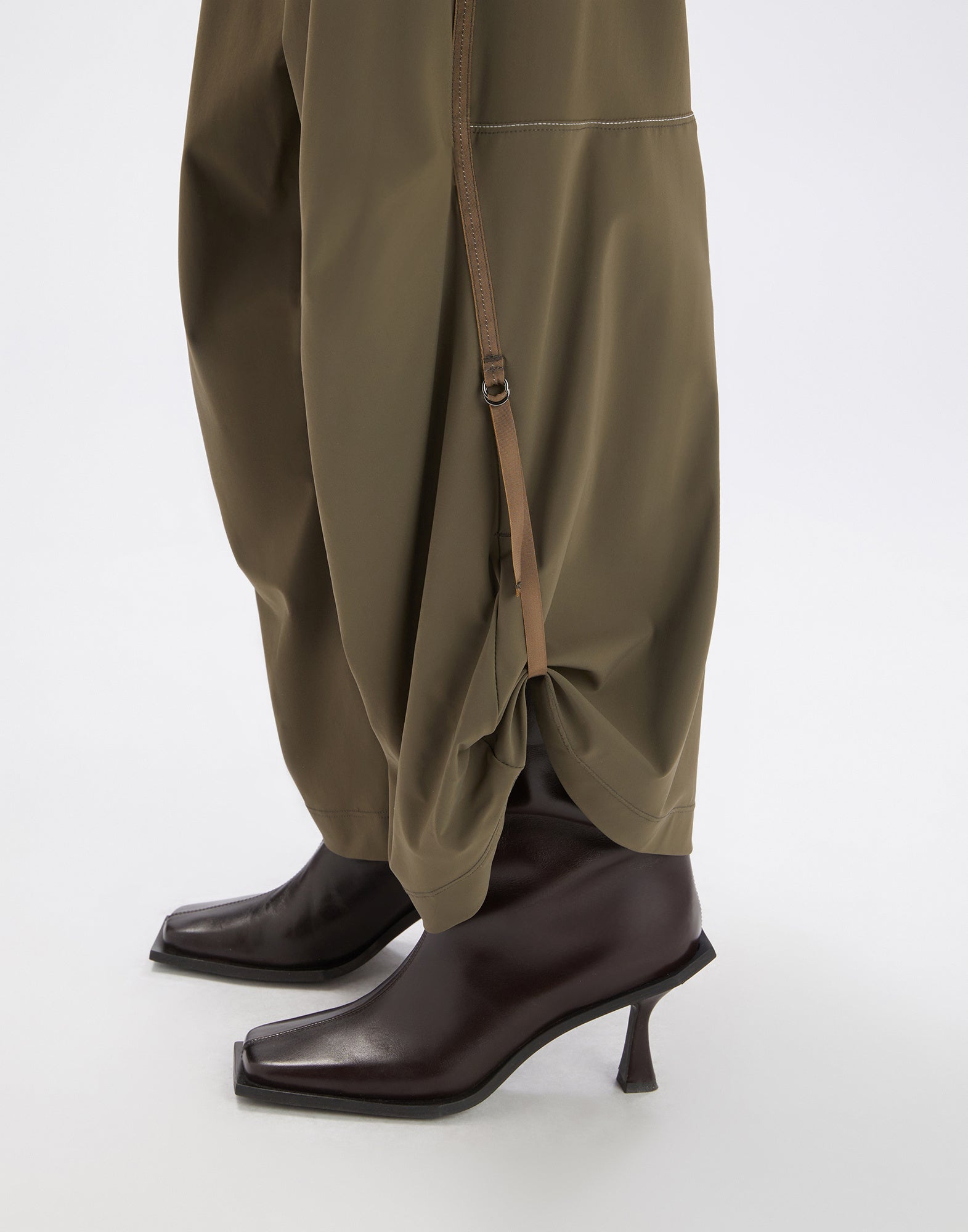 Side view of the OUTGOING trousers, olive green wide legs gathered at the hem with strap and D-ring, worn over brown ankle boots.