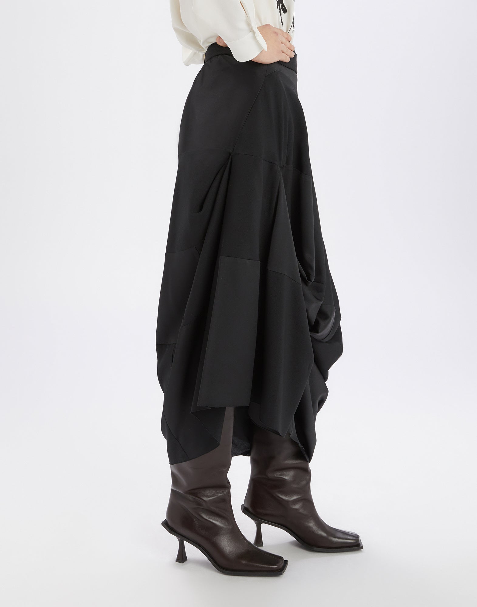 Side view of the GEEK skirt in black flowing tech satin with asymmetric draping, midi length, and elastic waistband, worn with boots.