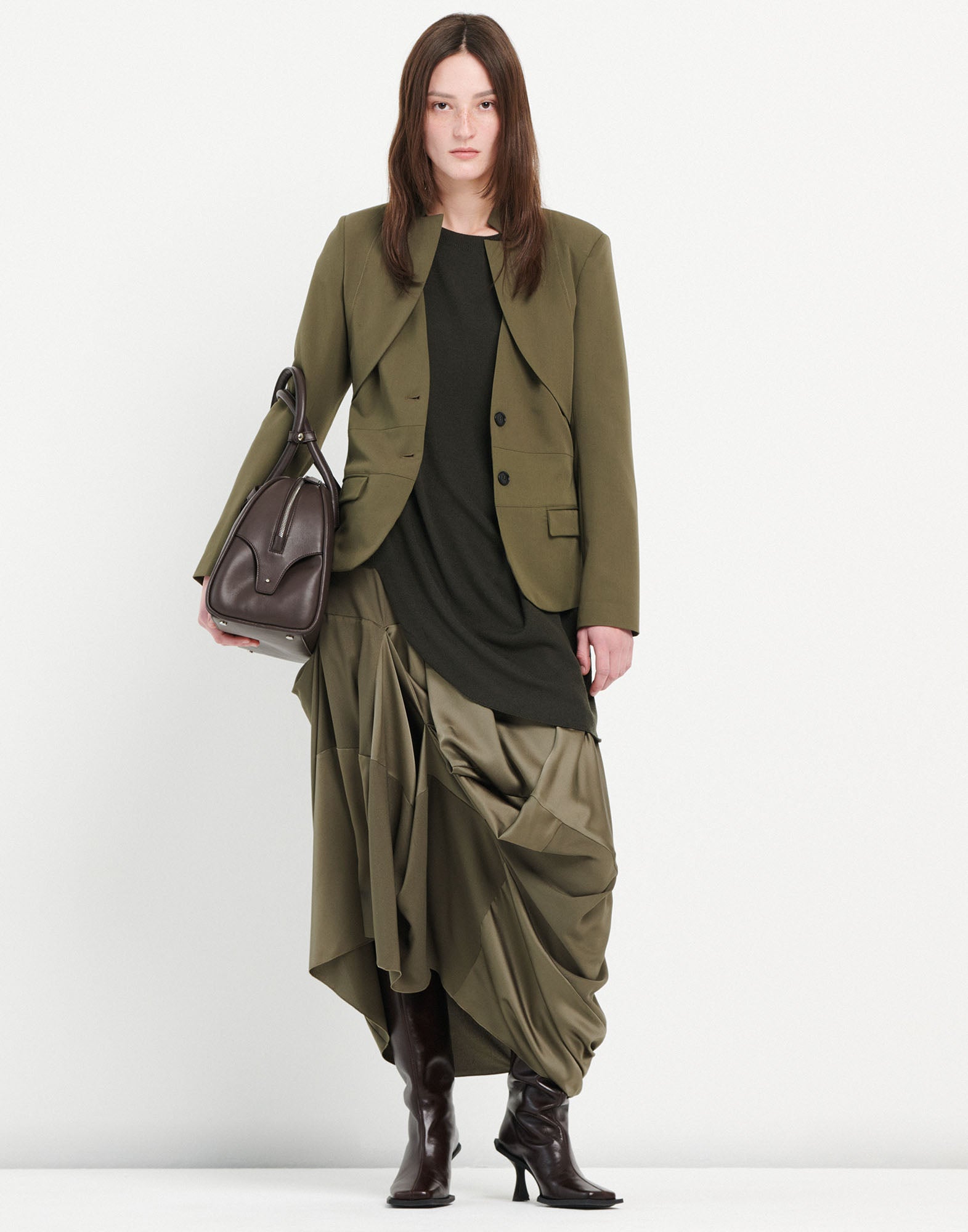 Front view of a woman wearing the GEEK skirt in fluid tech satin with asymmetric draping, midi length, and elastic waistband. Styled with blazer, boots, and bag.