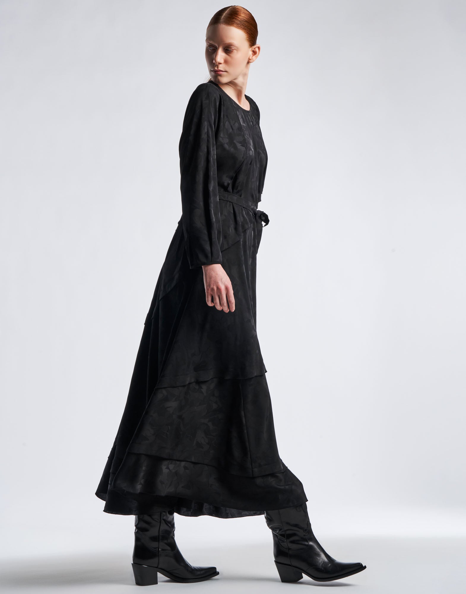 Seitenansicht einer Frau im FAVOURITE Maxi-Kleid aus schwarzem Funktionssatin mit Damast-Blumenmuster, langen Dolmanärmeln und weitem, mehrlagigem Rock.