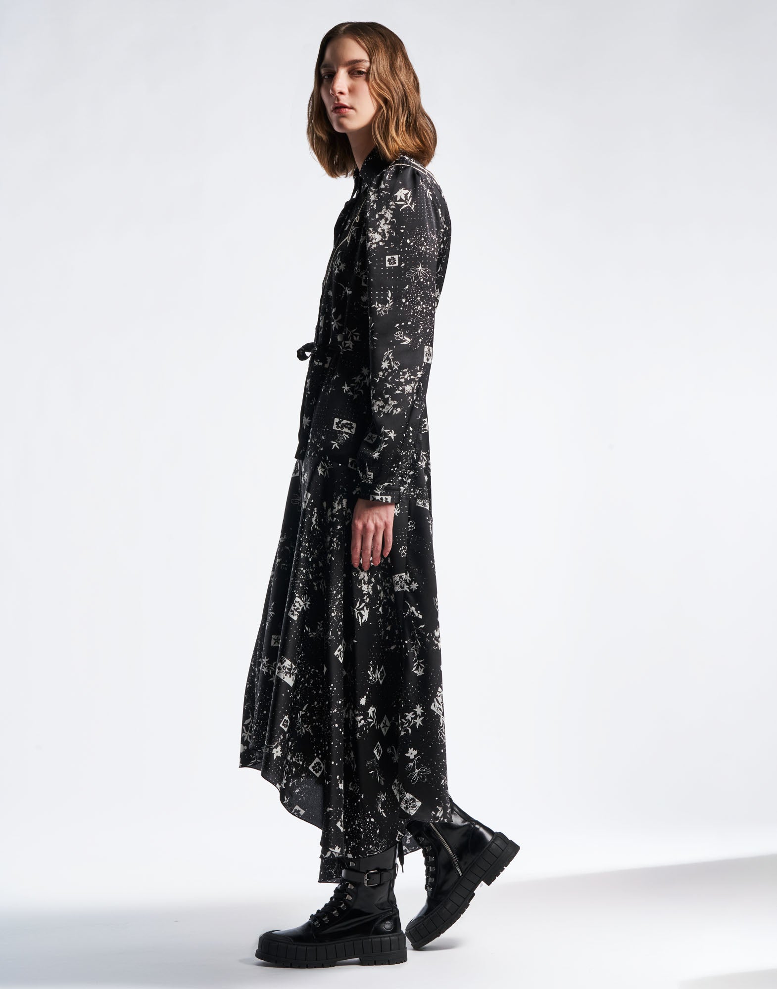 Side view: Model wears the COMPELLING shirtdress in black technical satin with geometric floral pattern and ivory splashes, full skirt, and lace-up boots.