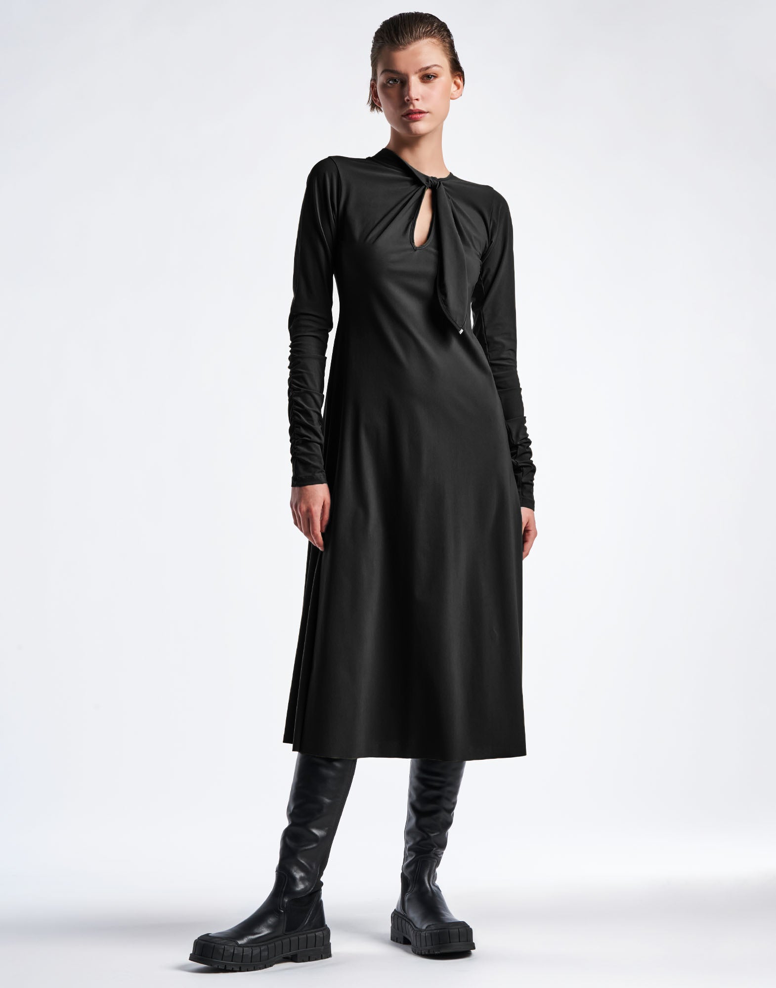 Frontal view: SINCERELY presents a flared black dress in Sensitive(R) sports jersey with long ruched sleeves, round neckline, self-fabric tie, and keyhole opening. The dress falls to mid-calf and is complemented by knee-high black boots.