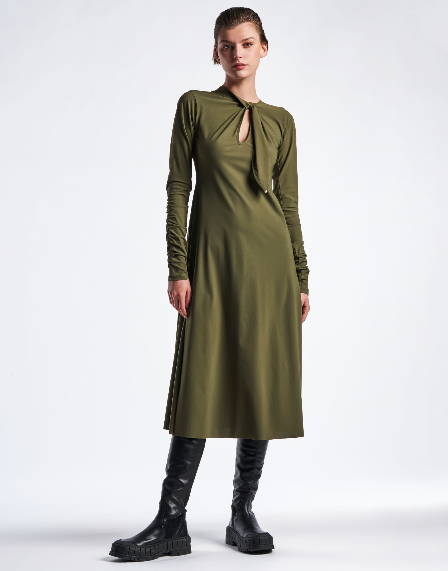 Frontal view: Model wears the SINCERELY dress in olive green Sensitive(R) sports jersey, featuring a round neckline with a tie and small slit at the front, long ruched sleeves, and flared hem.