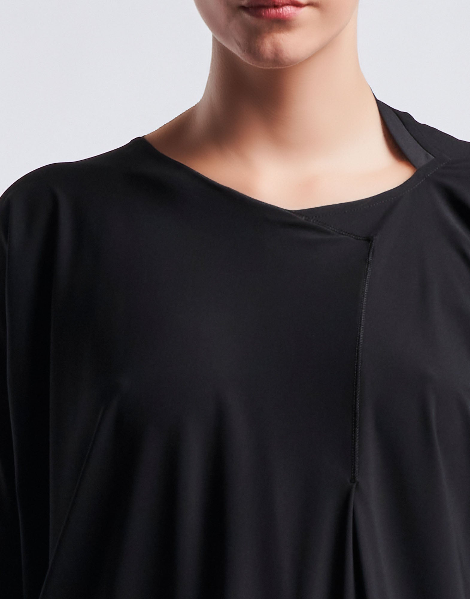 Detailansicht, Frontalansicht: PRELUDE Kleid zeigt den oberen Bereich mit asymmetrischem Ausschnitt, diagonaler Naht und feinem, glattem schwarzen Jersey-Stoff.