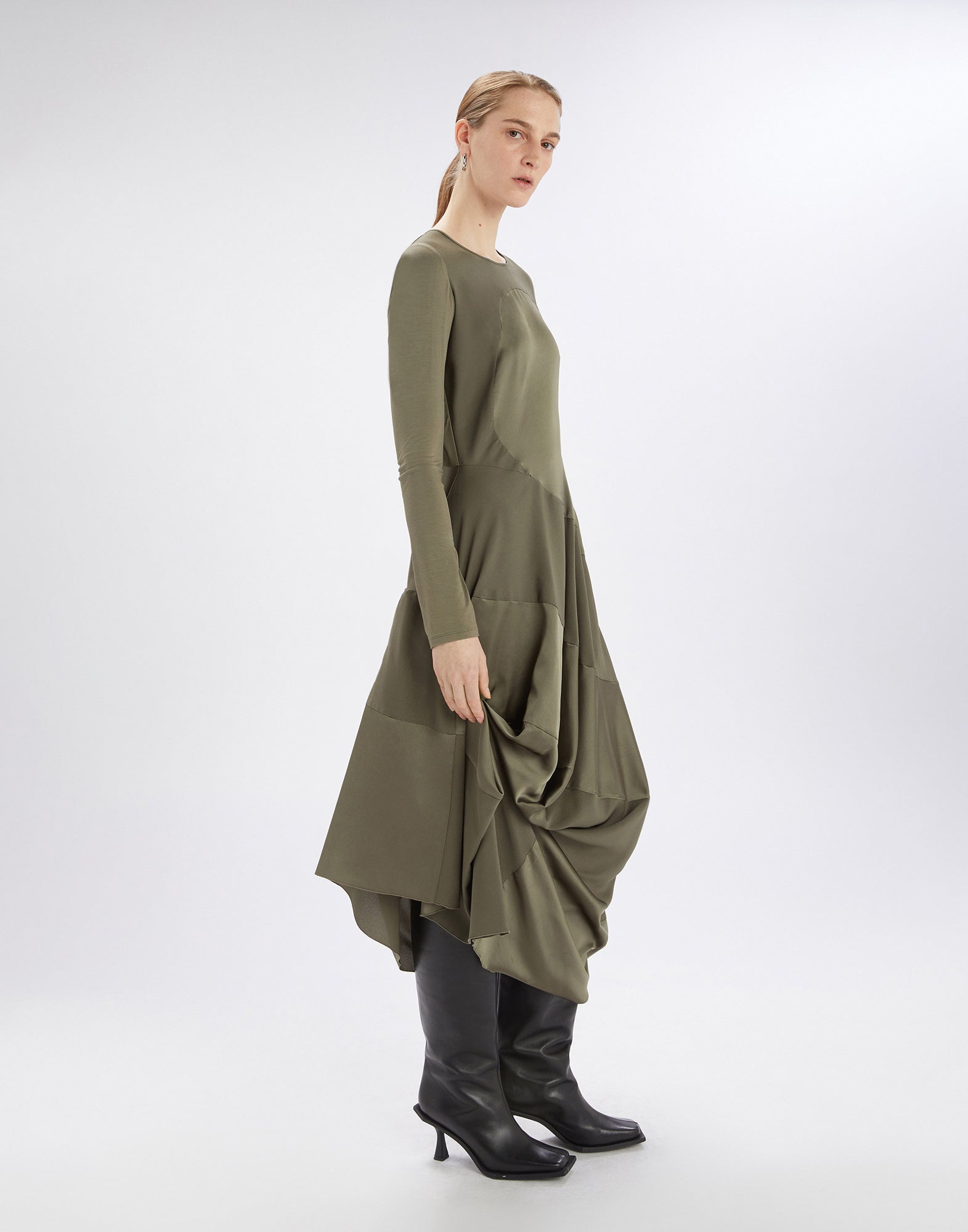 Side view of the PLACID dress worn by a model. The knee-length dress combines stretch jersey on the upper part with a satin-draped skirt in olive green. Long sleeves, round neckline, and black boots complete the look.