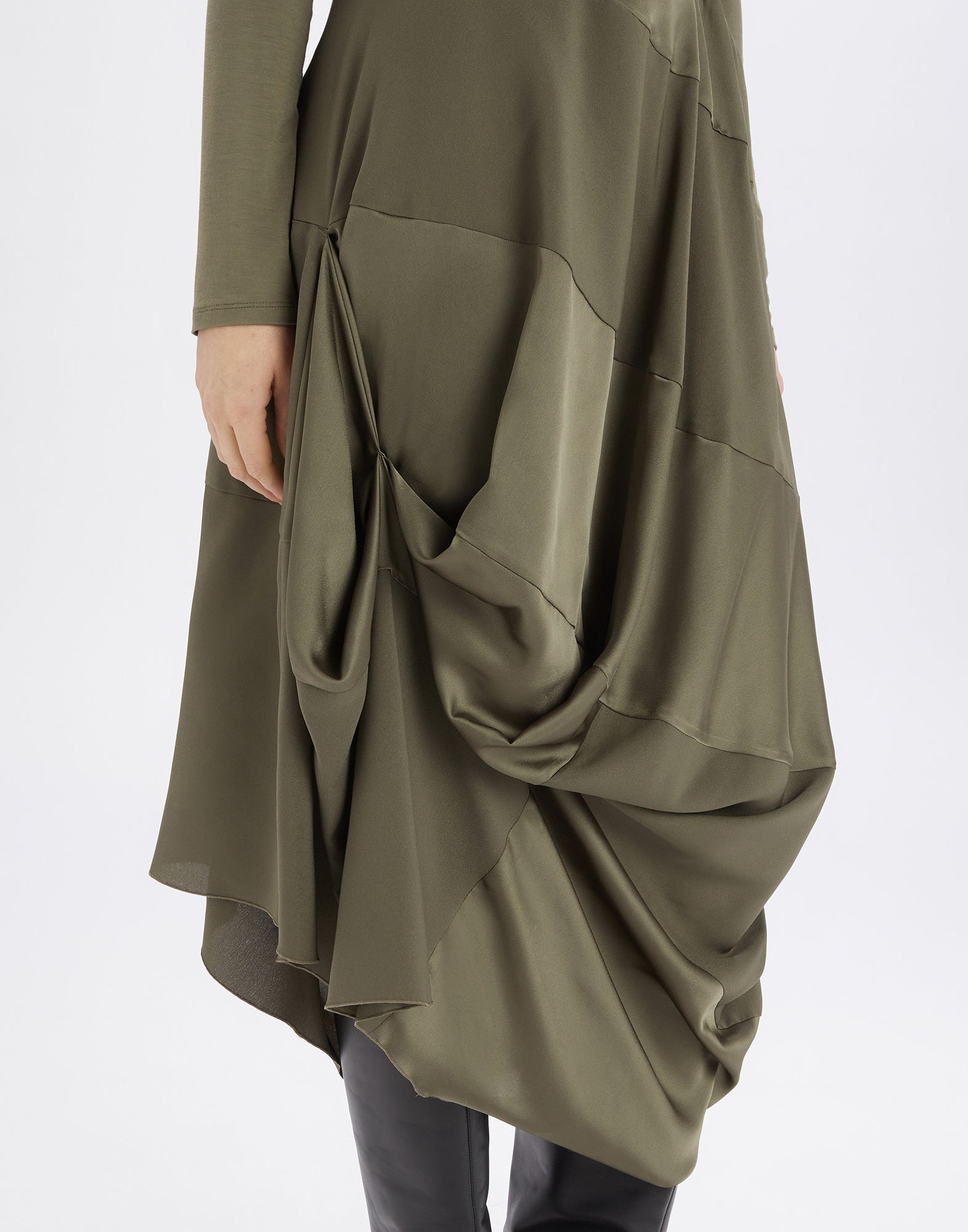 Close-up: lower part of the PLACID dress with draped olive green polyester satin, visible seam and pleat lines, a hand holding the hem.