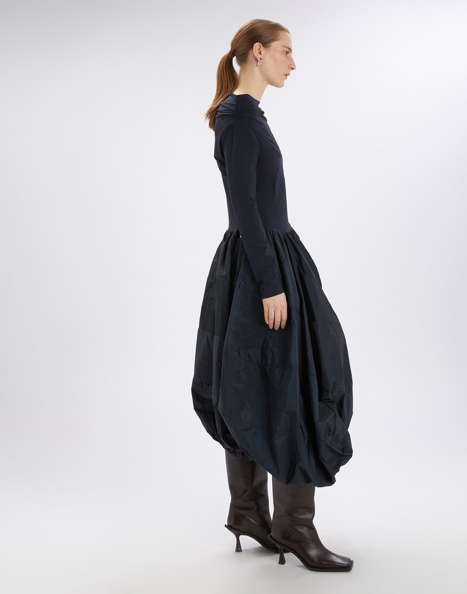 Seitenansicht des GRID Kleides, getragen von einer Frau, mit schlichtem, körpernahem Oberteil und ballonförmigem Rock aus glänzendem Taft, kombiniert mit langen Ärmeln und kniehohen Stiefeln.