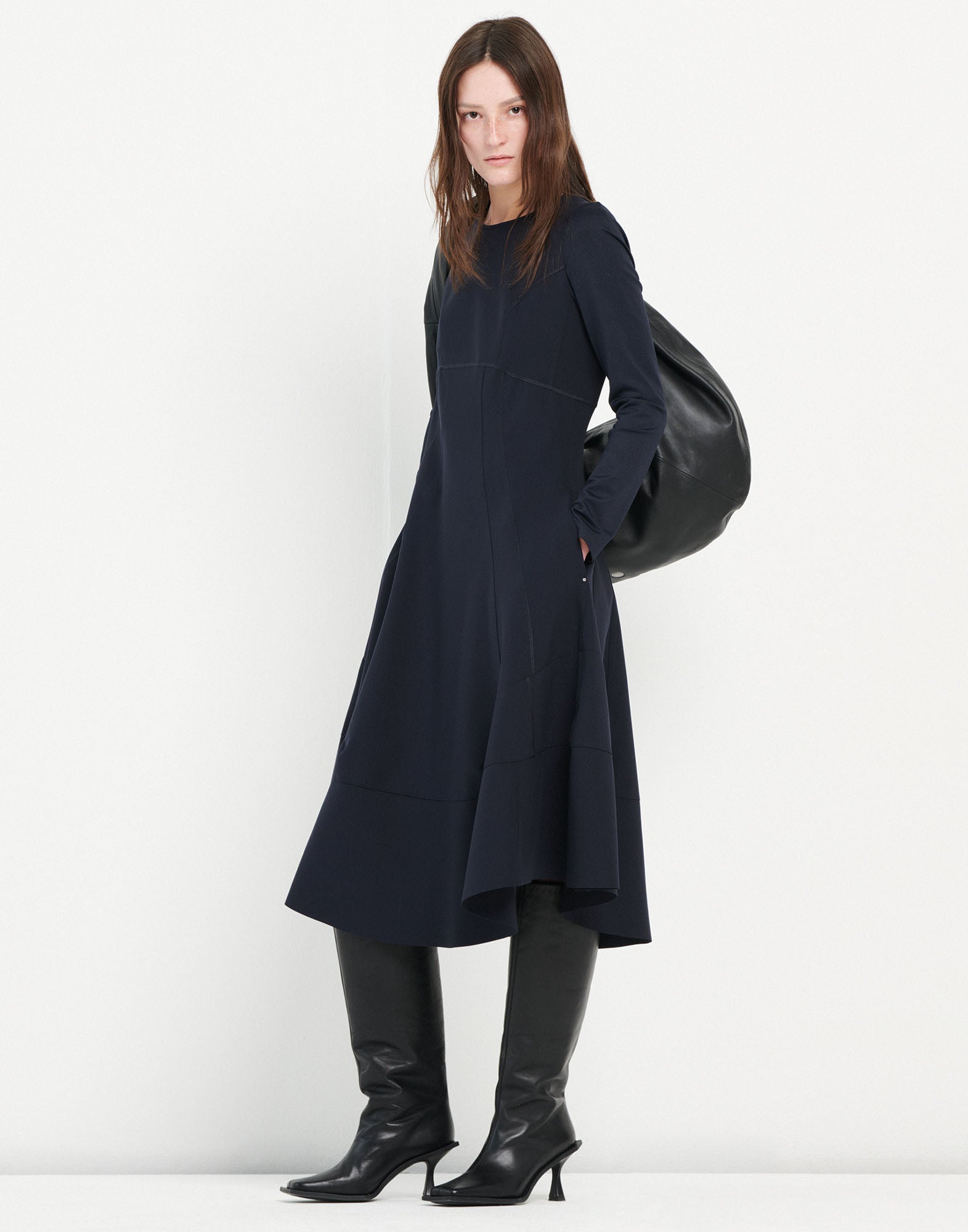 Frontalansicht: Modell trägt das Kleid SYNCHRO in Dunkelblau, langärmlig mit rundem Ausschnitt und asymmetrischem Saum, dazu kniehohe schwarze Stiefel und großem schwarzen Beutel.