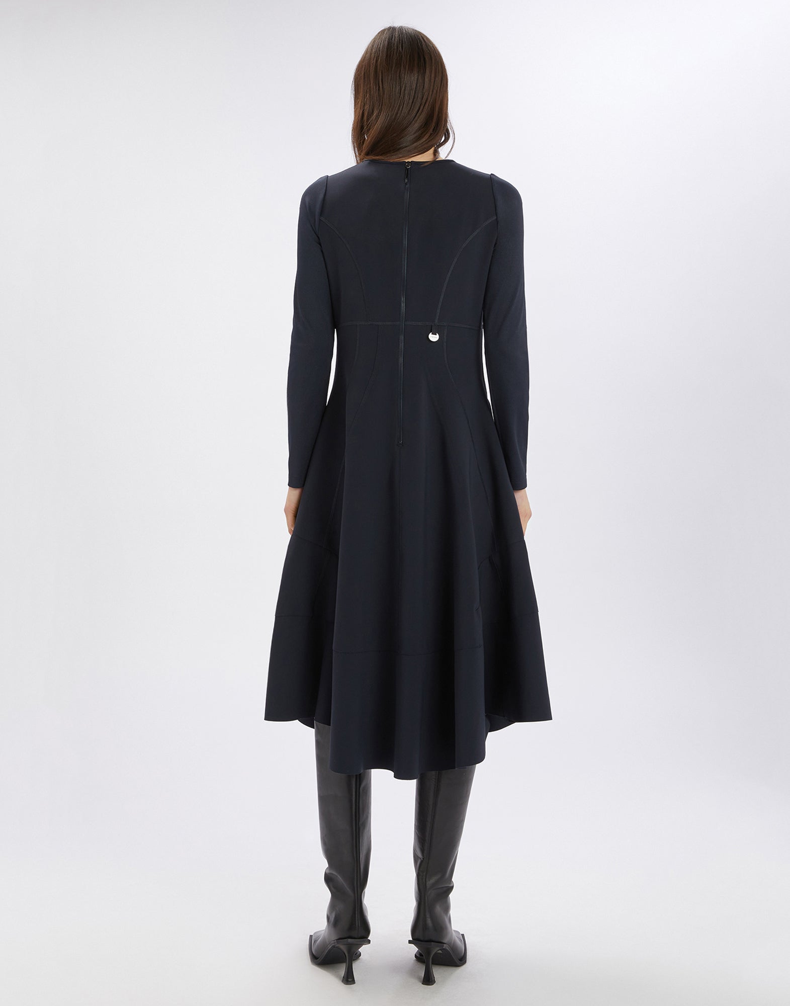 Rückansicht des SYNCHRO Kleids, Figur steht aufrecht, zeigt langen Ärmel, sichtbare Nähte und Reißverschluss am Rücken, Saum asymmetrisch, kniehohe Stiefel.