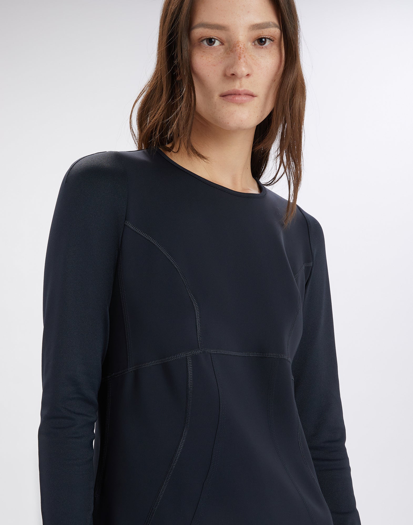 Frontalansicht einer Frau, die das Kleid SYNCHRO trägt: minimalistisches, schwarzes Langarmkleid mit sichtbaren Steppnähten, rundem Ausschnitt und betonter Taille.