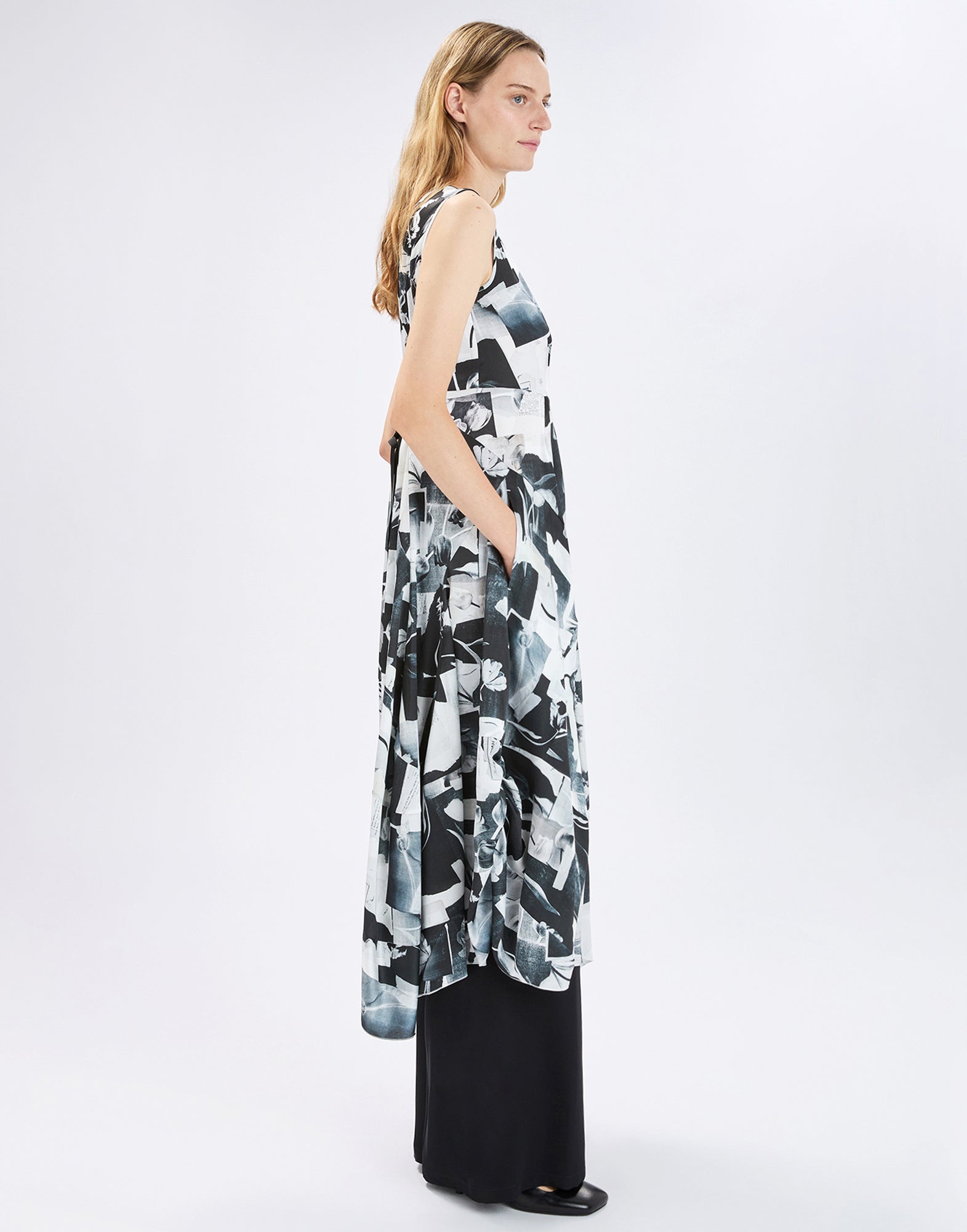 Side view: model wears the sleeveless SPARKY dress with black-and-white “Polaroid Tulips” print, wide asymmetric skirt over long black trousers, hands resting casually in the side pockets.