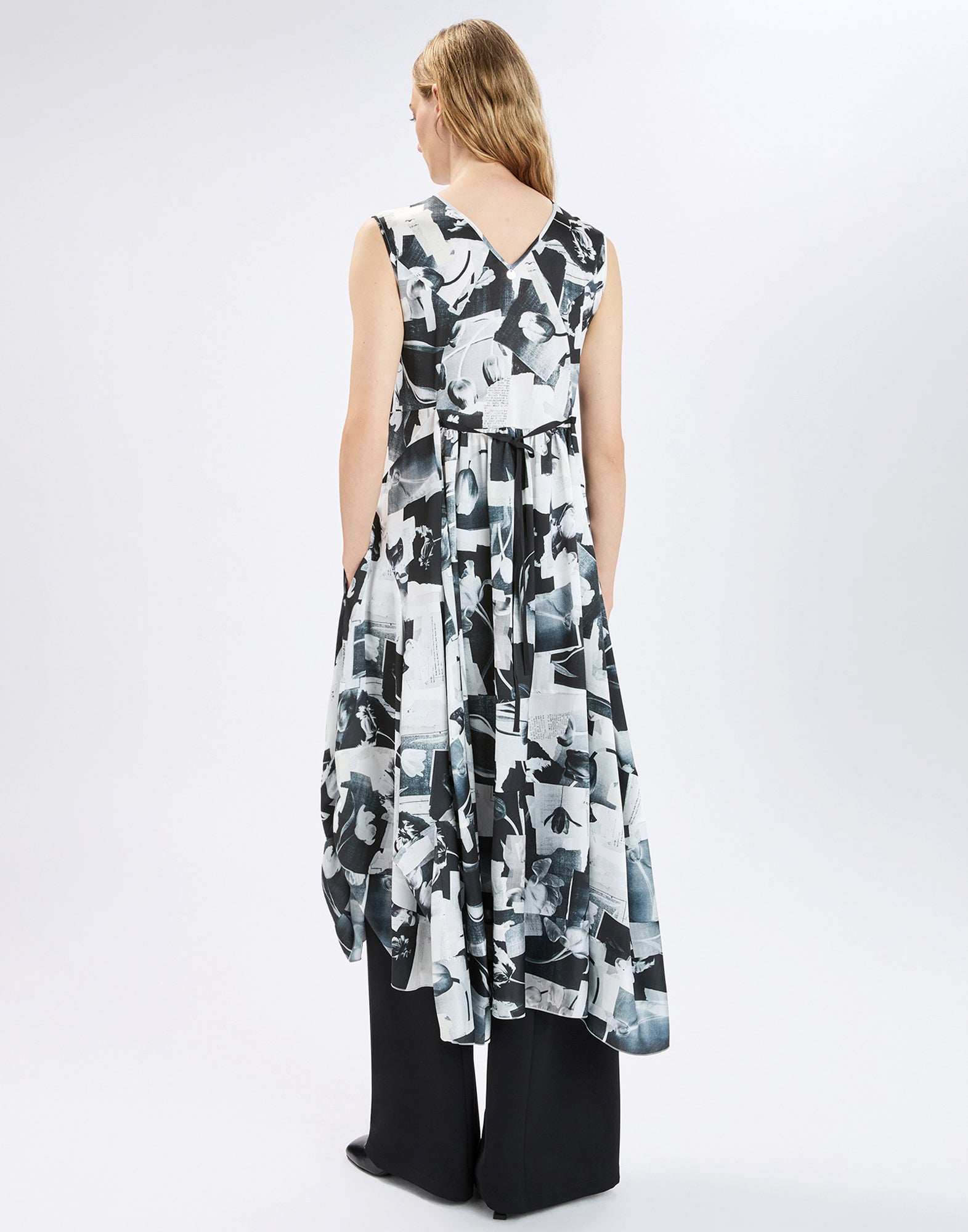 Back view: model wears sleeveless black-and-white patterned SPARKY dress with V-neckline at the back and loosely draped asymmetric hem over wide black trousers against a light studio background.
