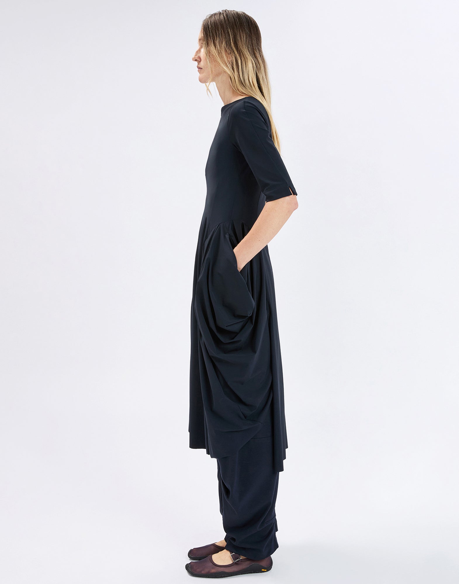 Side view: person wearing the dark, ankle-length NEW MESSENGER dress with boat neckline, elbow-length sleeves, draped left side with pocket, and asymmetrical hem that is longer in the back.