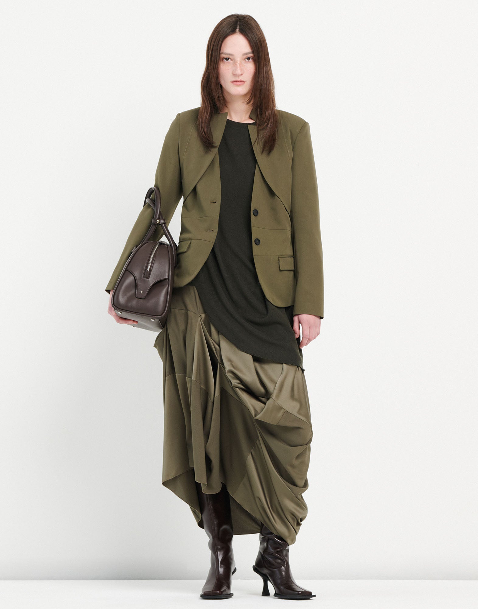 Front view of a woman wearing the PARALLAX jacket: olive green, textured blazer with inverted lapel, geometric neckline, button closure, and flap pockets.