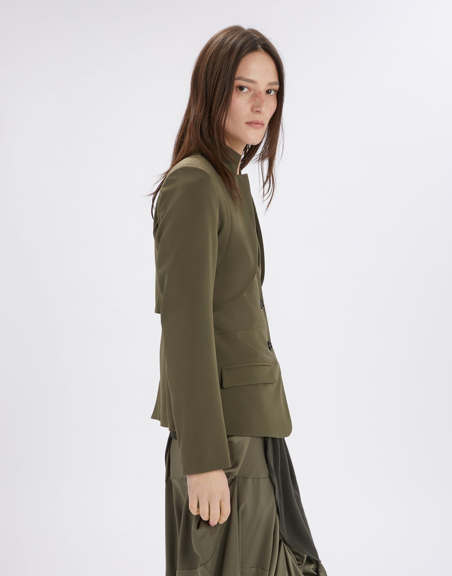 Side view of a person wearing the PARALLAX jacket in olive green; the modern design with inverted notched collar and exposed seams is clearly visible.
