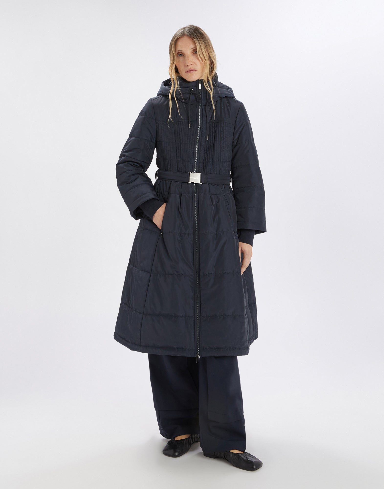 Front view: A woman wears the long, dark blue SCUTTLE down jacket with stand collar, hood, zipper, elastic waist belt, and quilted bodice.