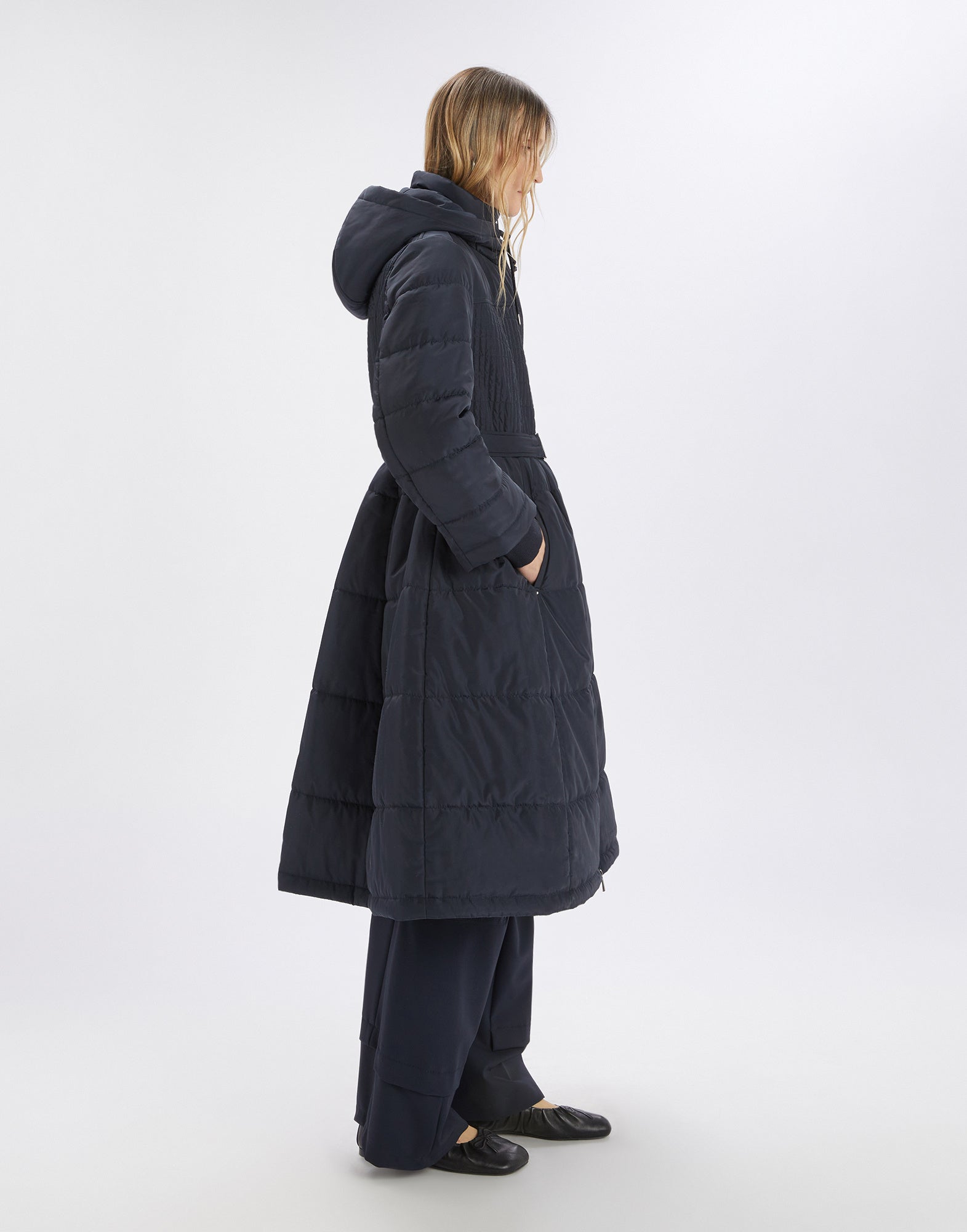 Side view of the SCUTTLE down jacket worn by a model. The jacket is dark, features a stand collar, hood, waist belt, and flared hem.