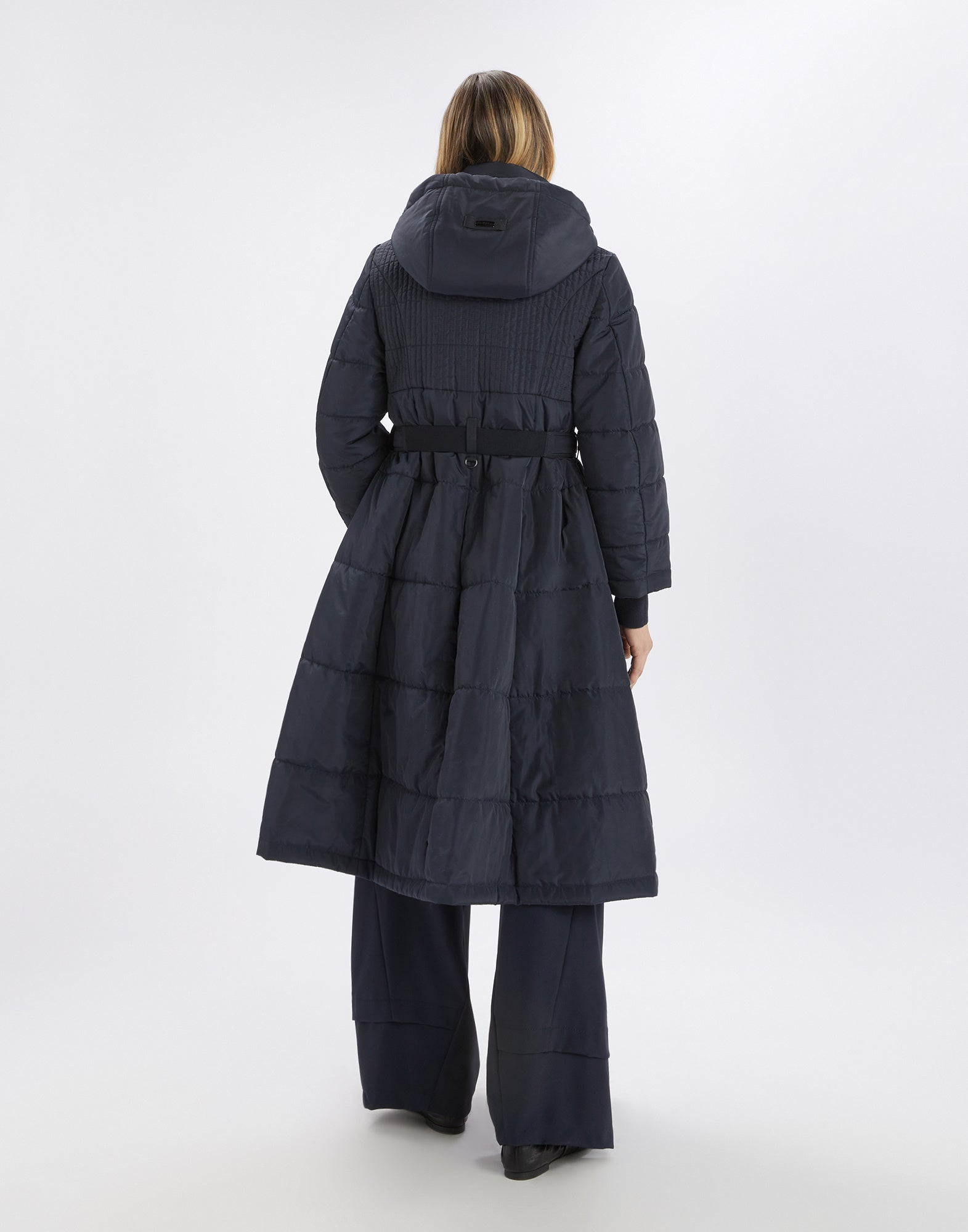 Back view of SCUTTLE down jacket: black, quilted down jacket with hood, elastic waist belt, and flared bell shape, worn by a woman.