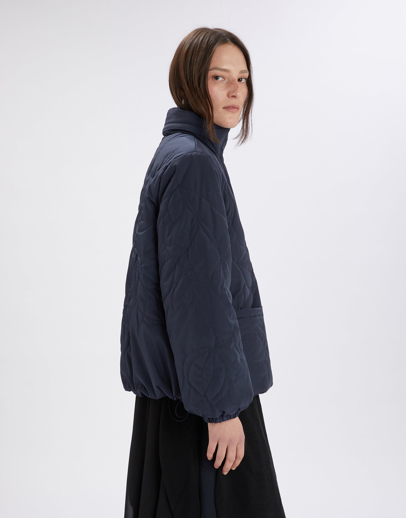 Seitenansicht einer Frau, die die BLURT Jacke trägt, welche aus dunkelblauem, gestepptem Nylon mit floraler Steppung, Stehkragen und aufgesetzten Taschen besteht.