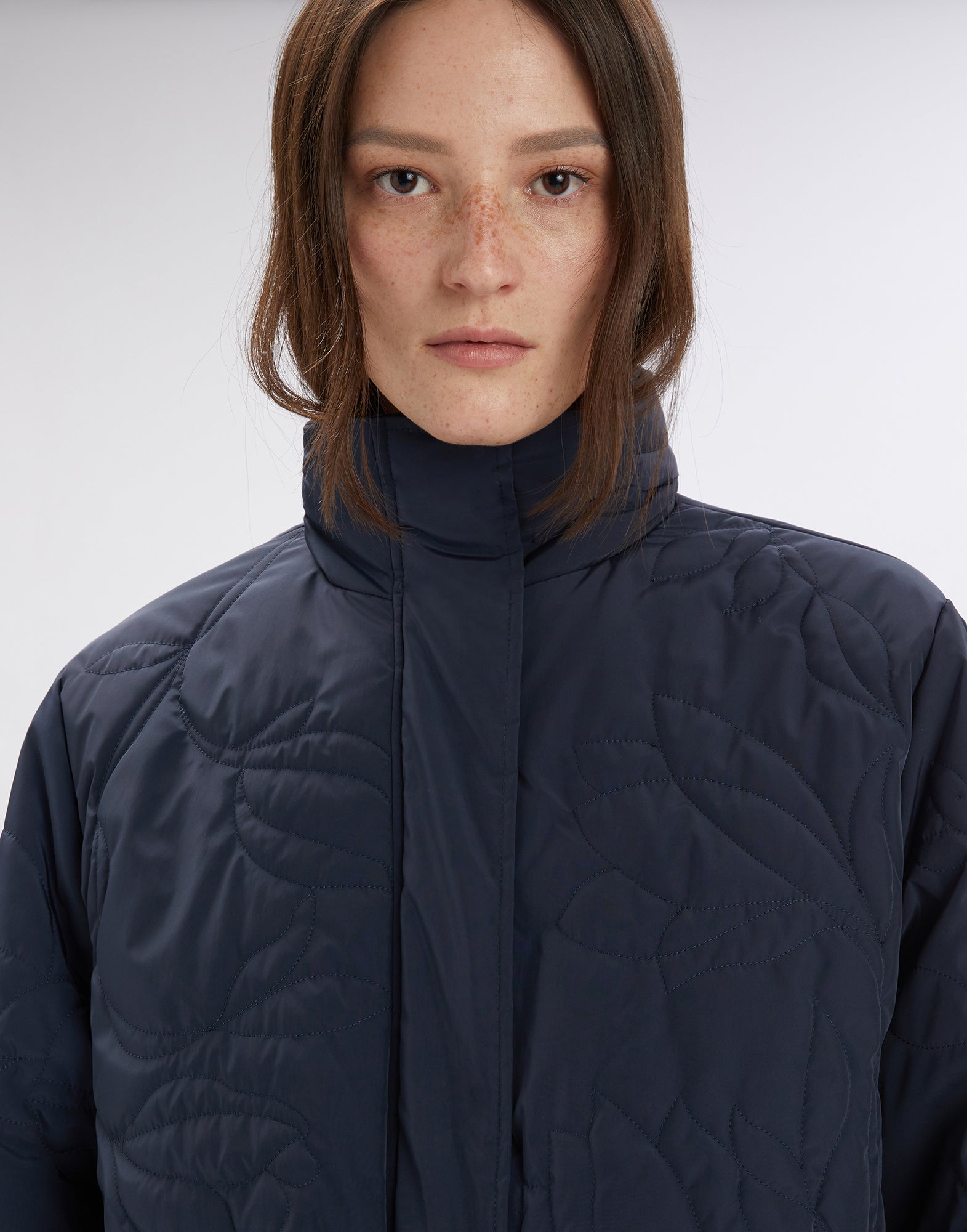 Frontalansicht einer Frau, die die BLURT Jacke trägt; sportliches Design in dunklem Nylon mit Stehkragen und floraler Steppung, Nahaufnahme vom Oberkörper.