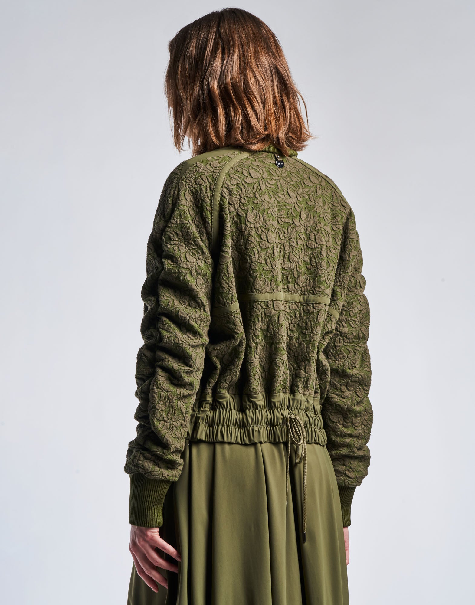 Rückansicht einer Person, die die BOAST Bomberjacke aus floral gemustertem, olivgrünem Jacquard-Baumwollmischjersey mit gerüschten Raglanärmeln und elastischem Taillenbund trägt.