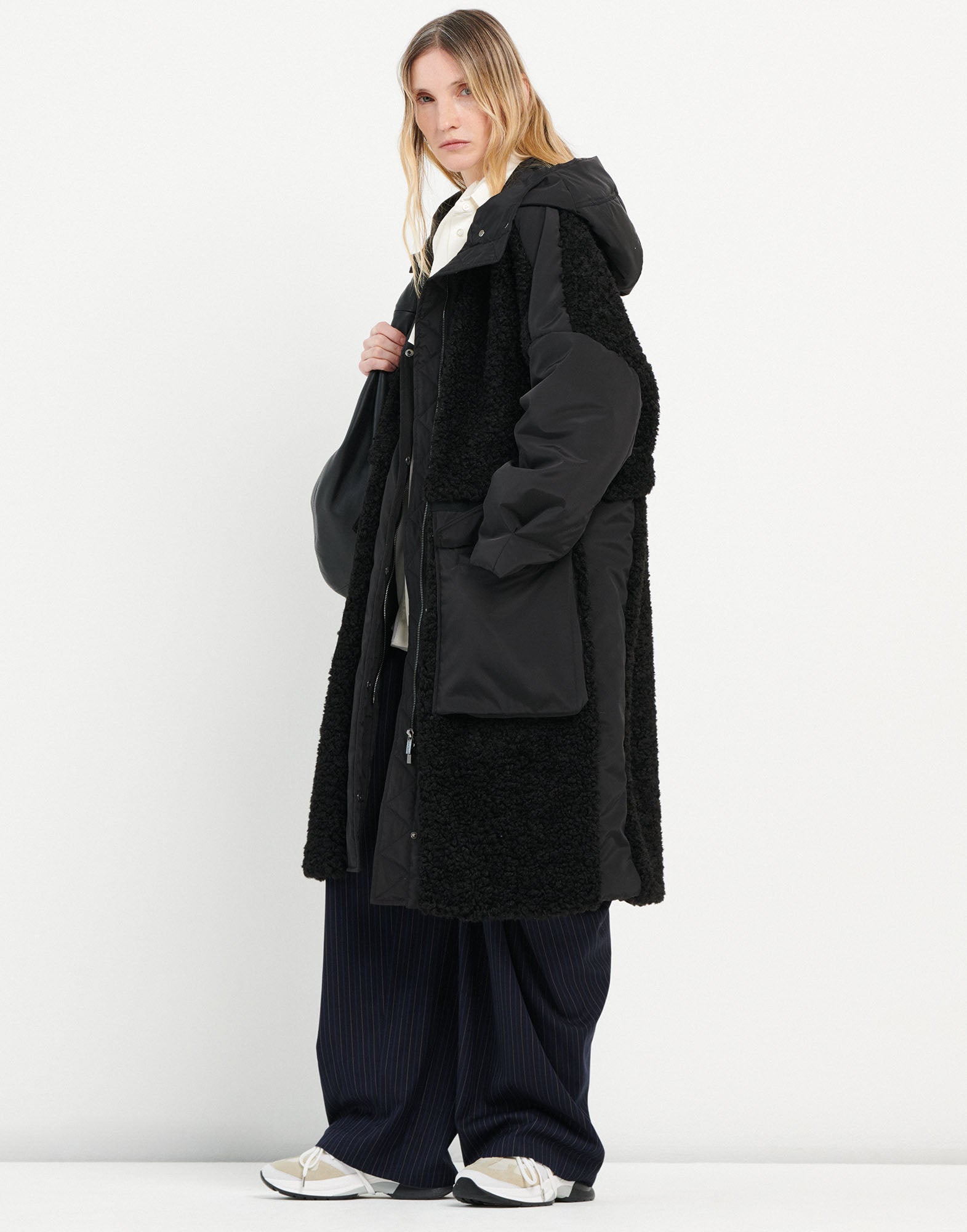Front view: A woman wears the ANNOTATE coat made of black sherpa fabric with nylon inserts, large hood, and pockets – sporty and knee-length.