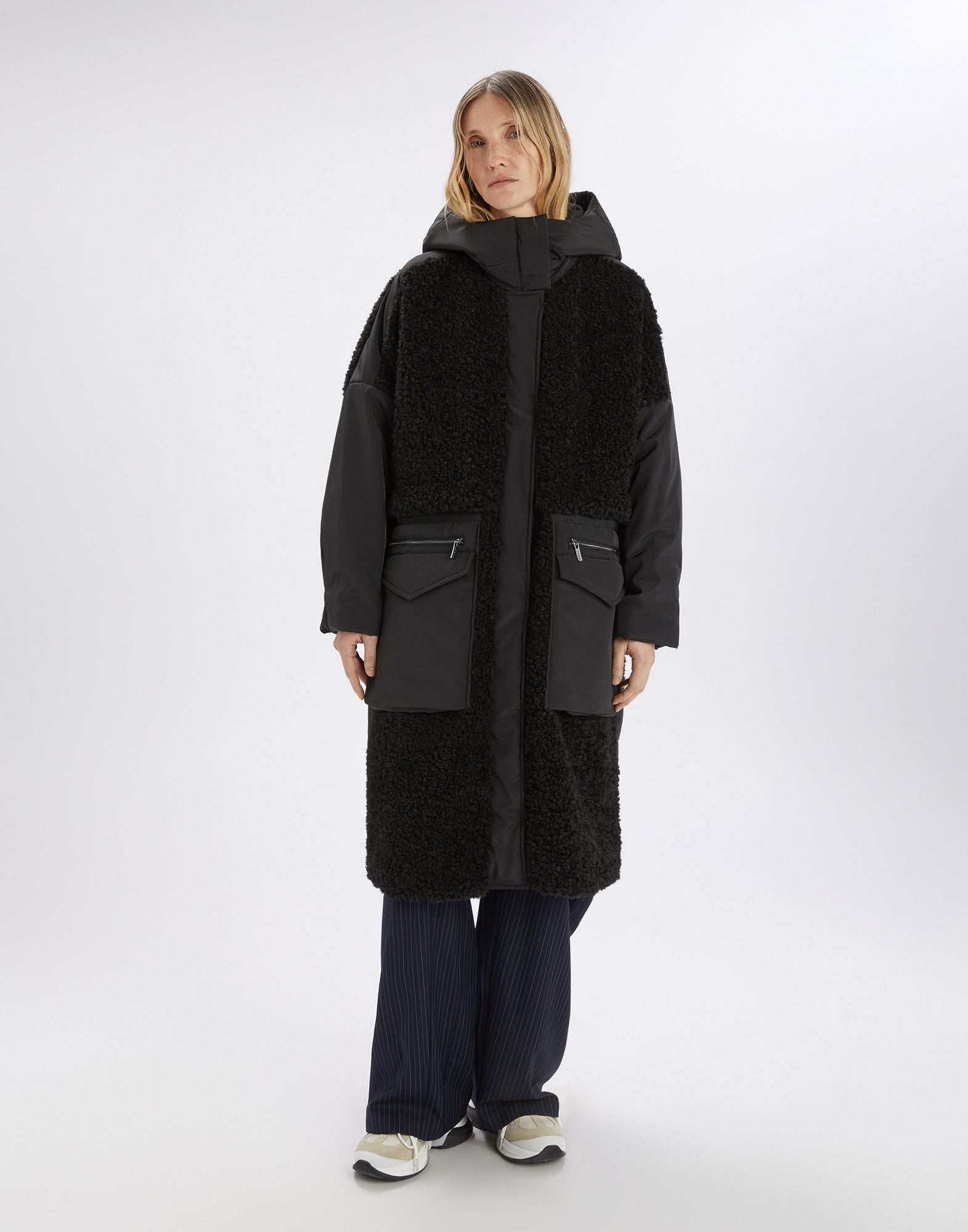 Front view: A woman wears the modern ANNOTATE coat in black with sherpa fabric, nylon inserts, hood, stand collar, and large front pockets.