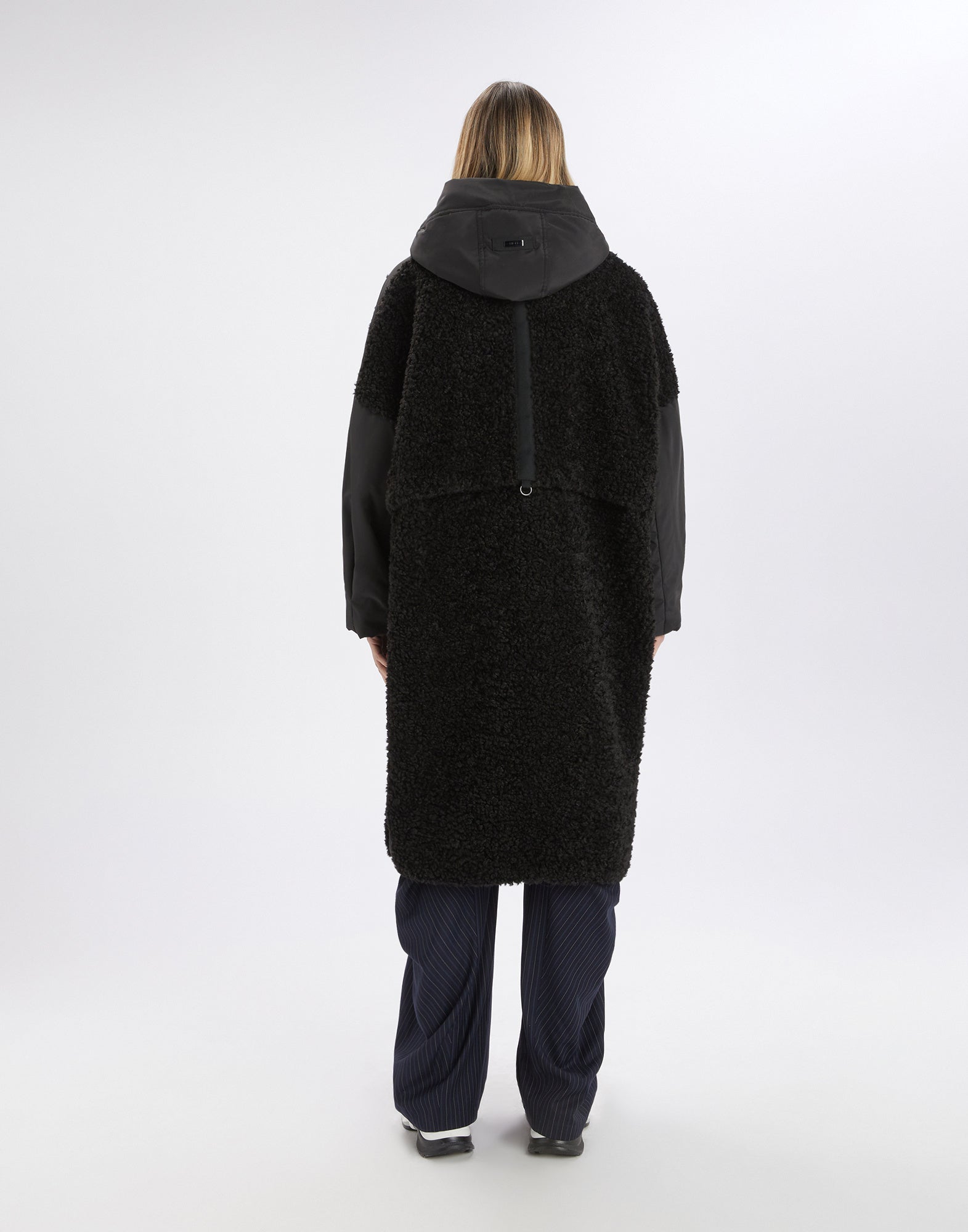 Rear view: person wears ANNOTATE coat with black sherpa fabric, nylon inserts, hood, and knee-length cut. The coat looks sporty and comfortable.