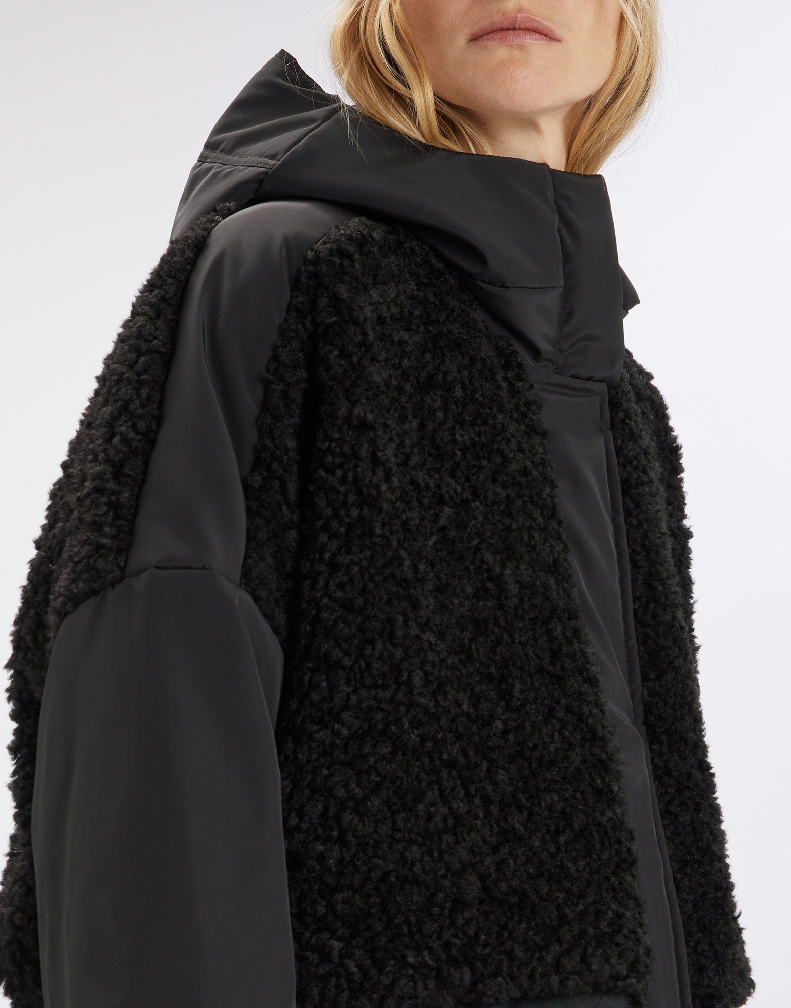 Detail view: woman wears ANNOTATE coat, black sherpa fabric with nylon inserts, hood and stand collar, soft, voluminous texture visible.