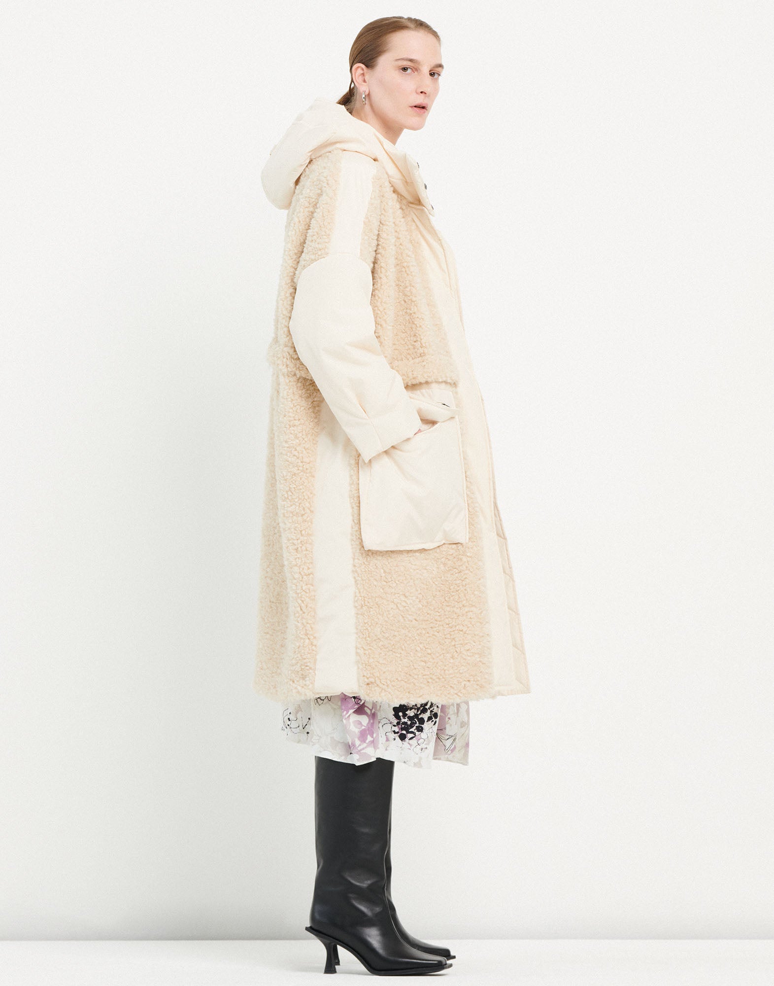 Side view of a person wearing the ANNOTATE coat. The knee-length coat is made of beige sherpa fabric with nylon inserts, hood, and large front pockets.