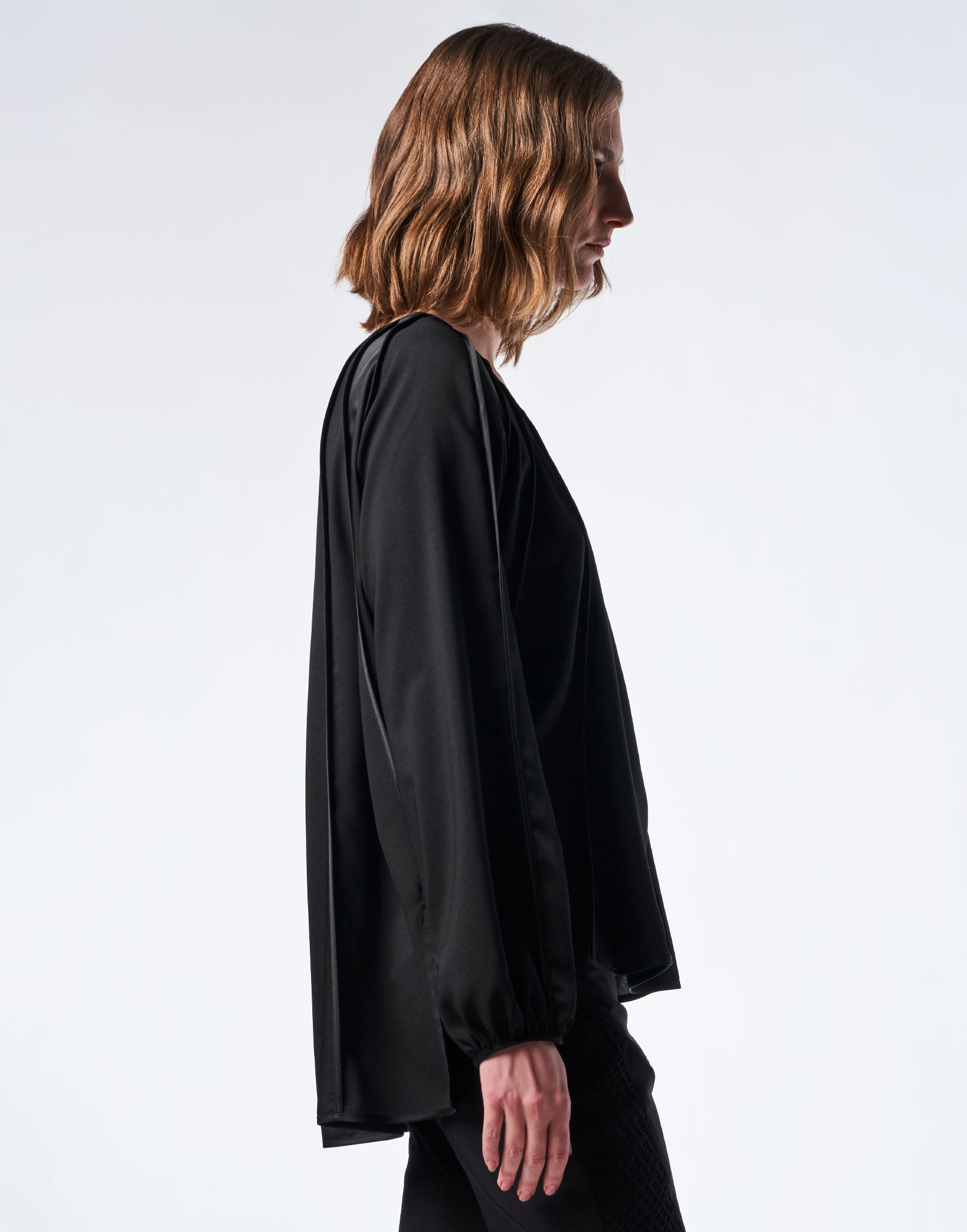 Seitenansicht einer Person, die das Oberteil GIVE CHASE trägt, mit lockerem Schnitt, abwechselnd mattem und glänzendem schwarzem Kreppsatin, langen Raglanärmeln und Manschetten.