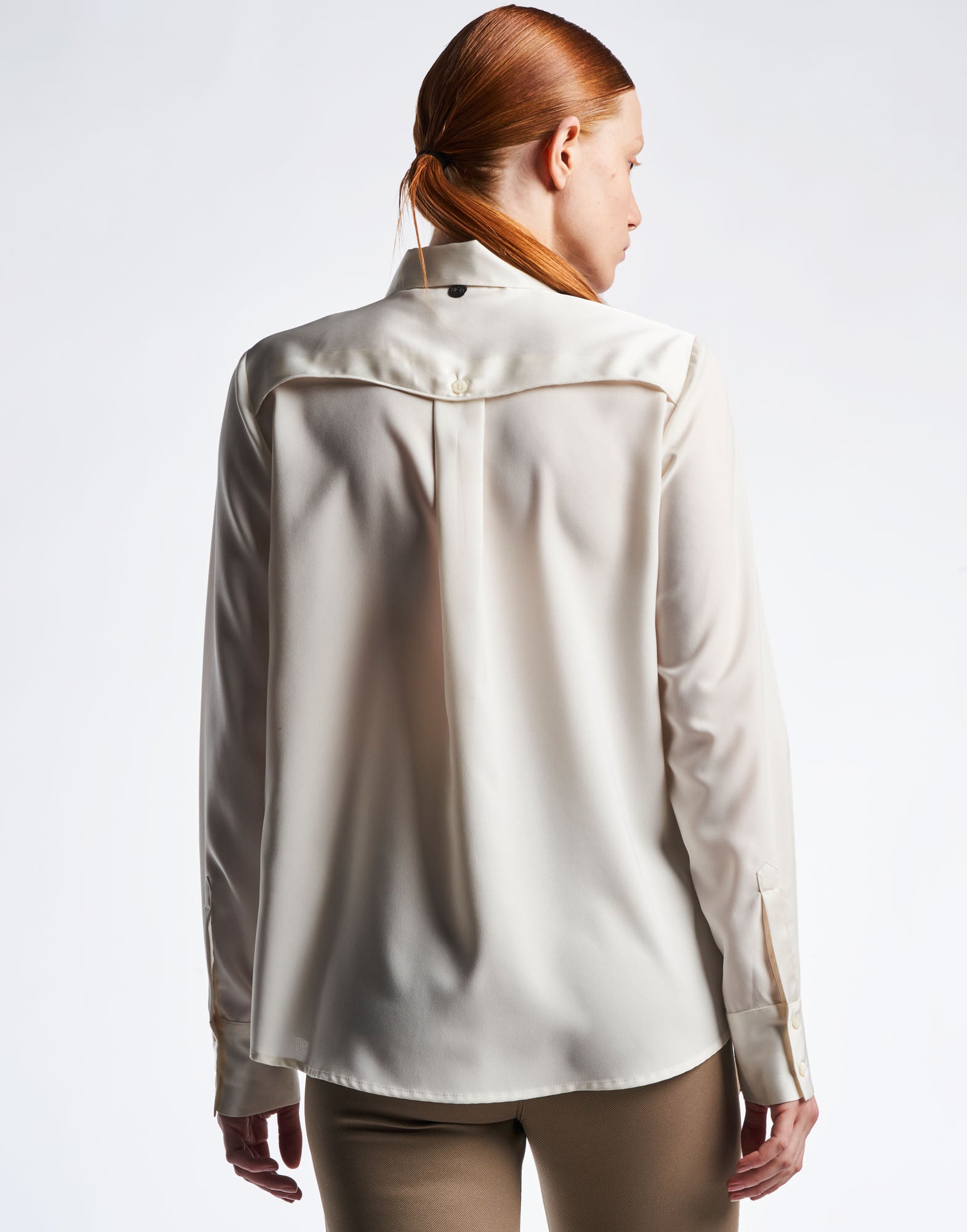 Rear view of a person wearing the ALTERNATE blouse. The blouse is ivory, features an oval yoke with decorative button, and a curved, longer back hem.
