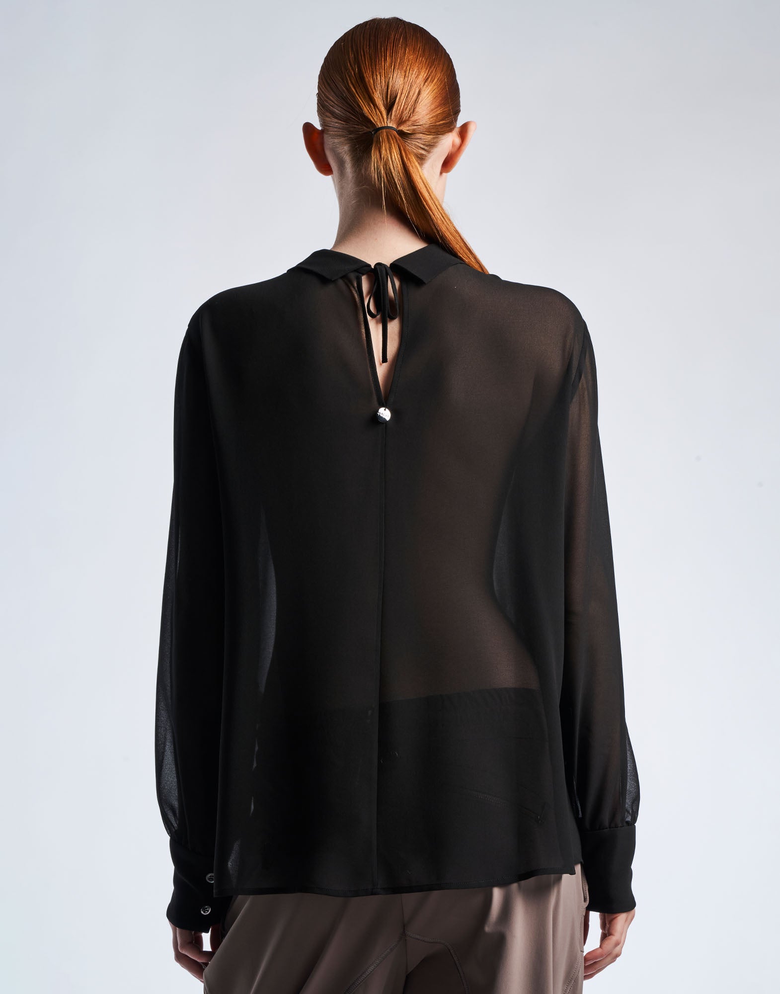 Rückansicht einer Person, die die VICE VERSA Bluse aus schwarzem, transparentem Funktions-Georgette mit V-Ausschnitt und Bindebändern am Nacken trägt.