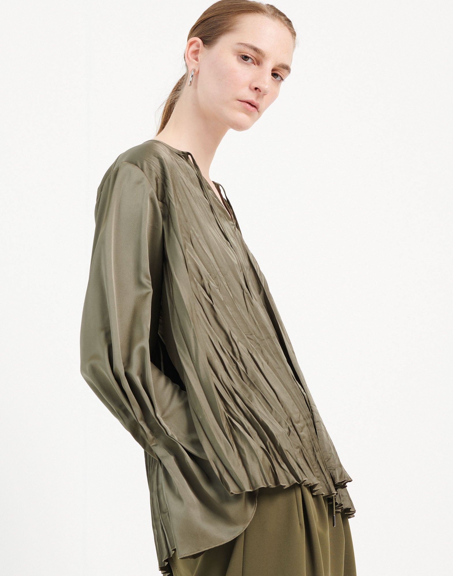 Side view of a woman wearing the ONTOP blouse; olive green shiny satin with pleated effect, wide long sleeves, and round neckline.