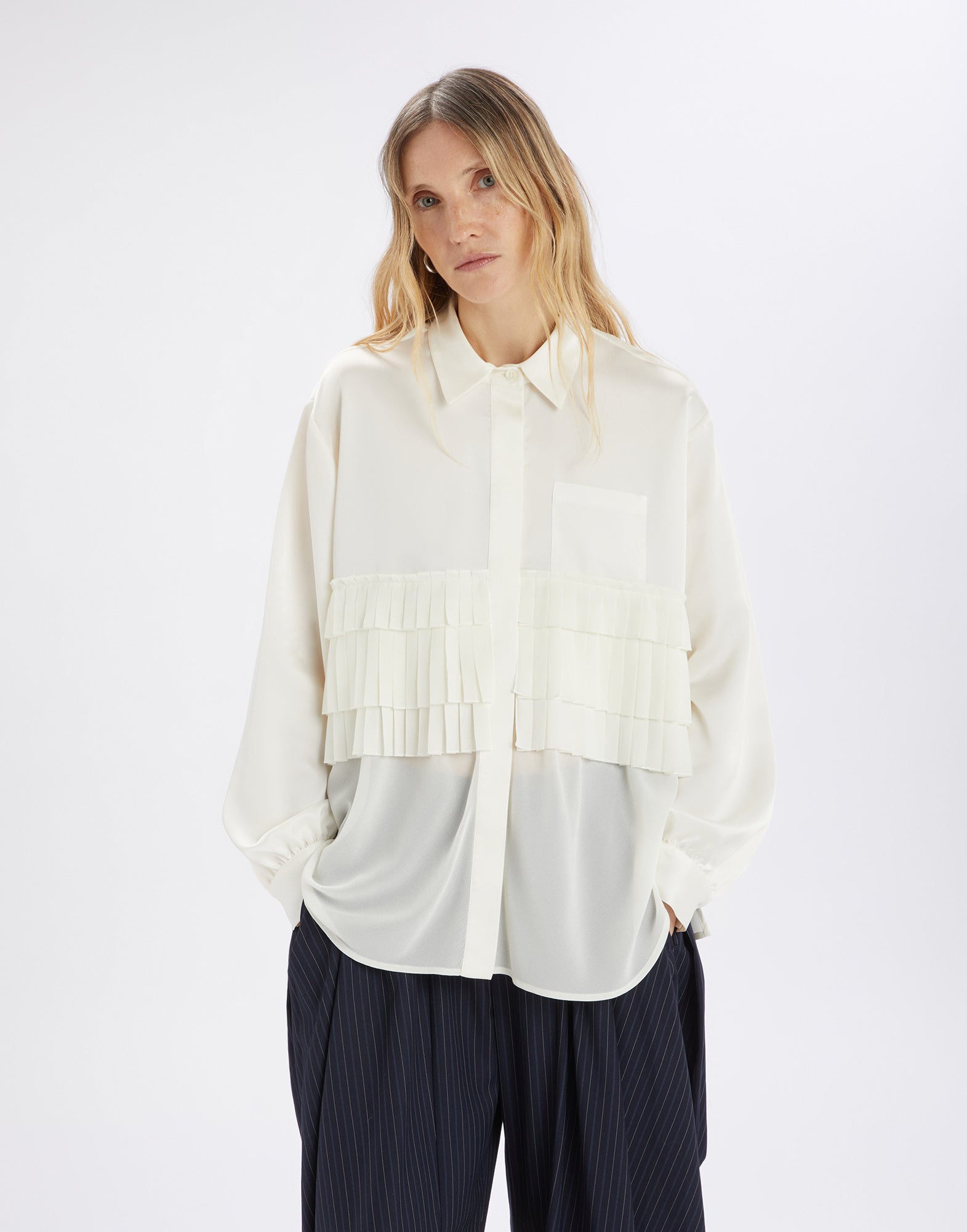 Front view of the MERYL blouse: woman wears a cream georgette blouse with three pleated ruffles at chest height, turn-down collar, concealed button placket, and rounded hem.