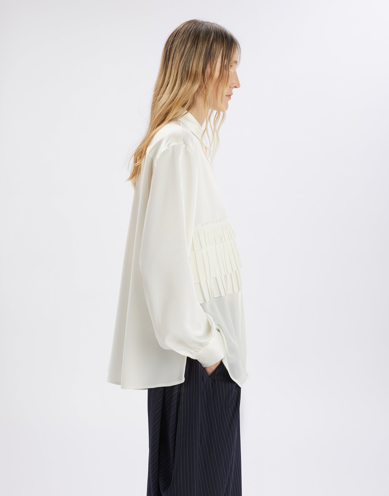 Side view of the MERYL shirt in cream white, worn by a person. The shirt features pleated ruffles on the upper body, long sleeves, and a loose fit.