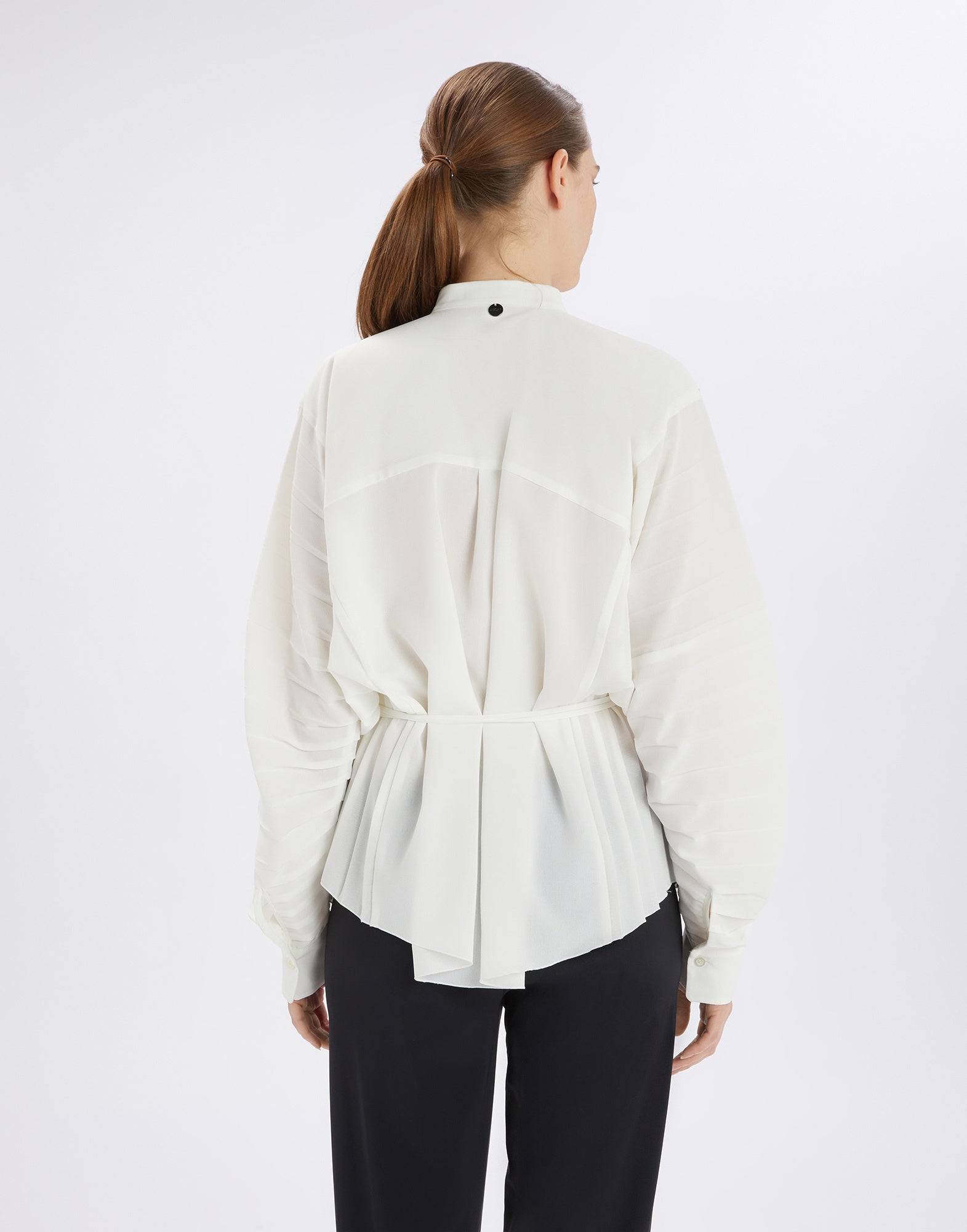 Back view of the white SLIM blouse with pleated details, mandarin collar, and waist drawstring, worn by a person with dark trousers.