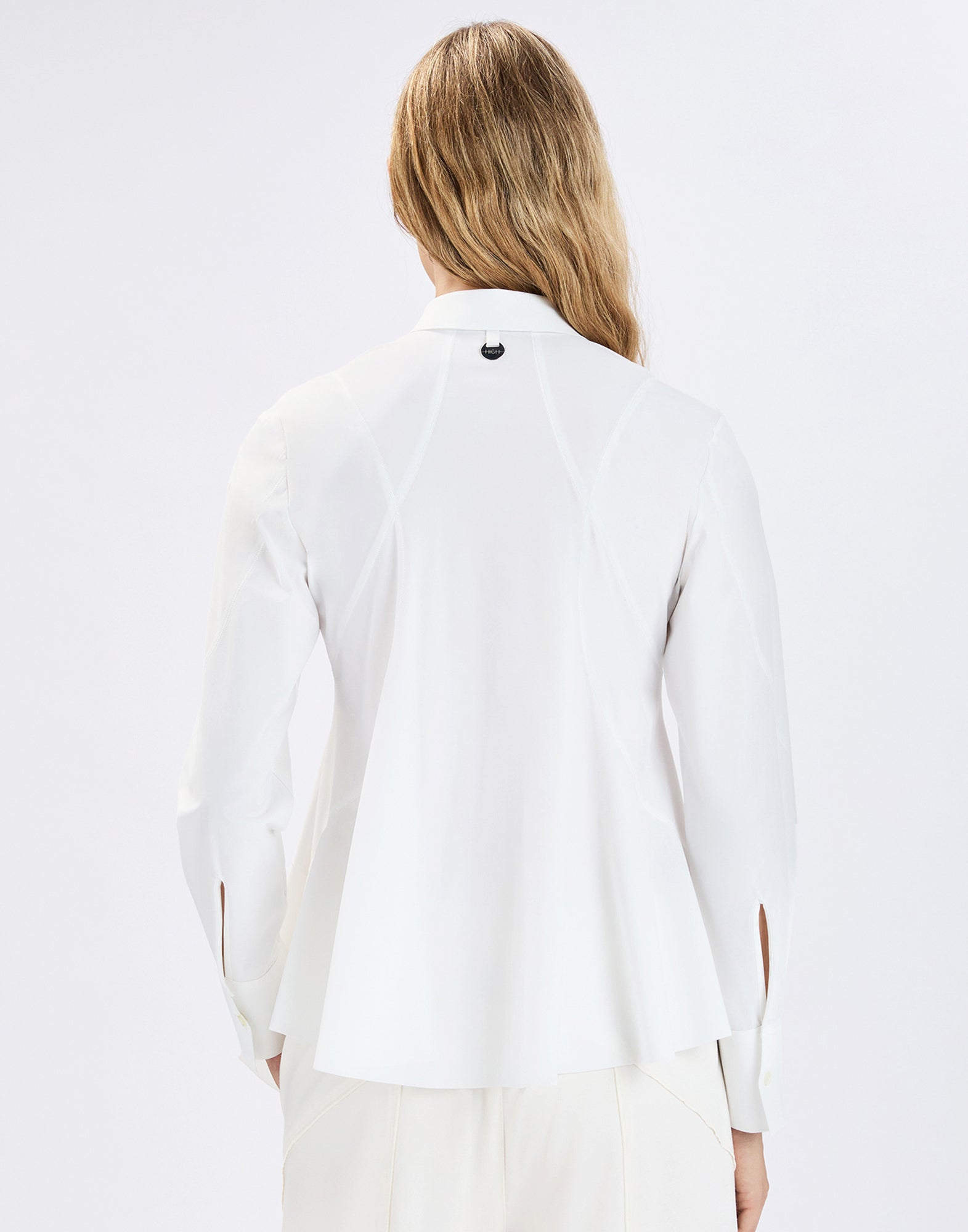 Rear view: Person wearing the white DEBONHAIR blouse with notched collar, visible shaping seams, loosely draped slightly asymmetrical hem, and long sleeves with slit and cuffs.
