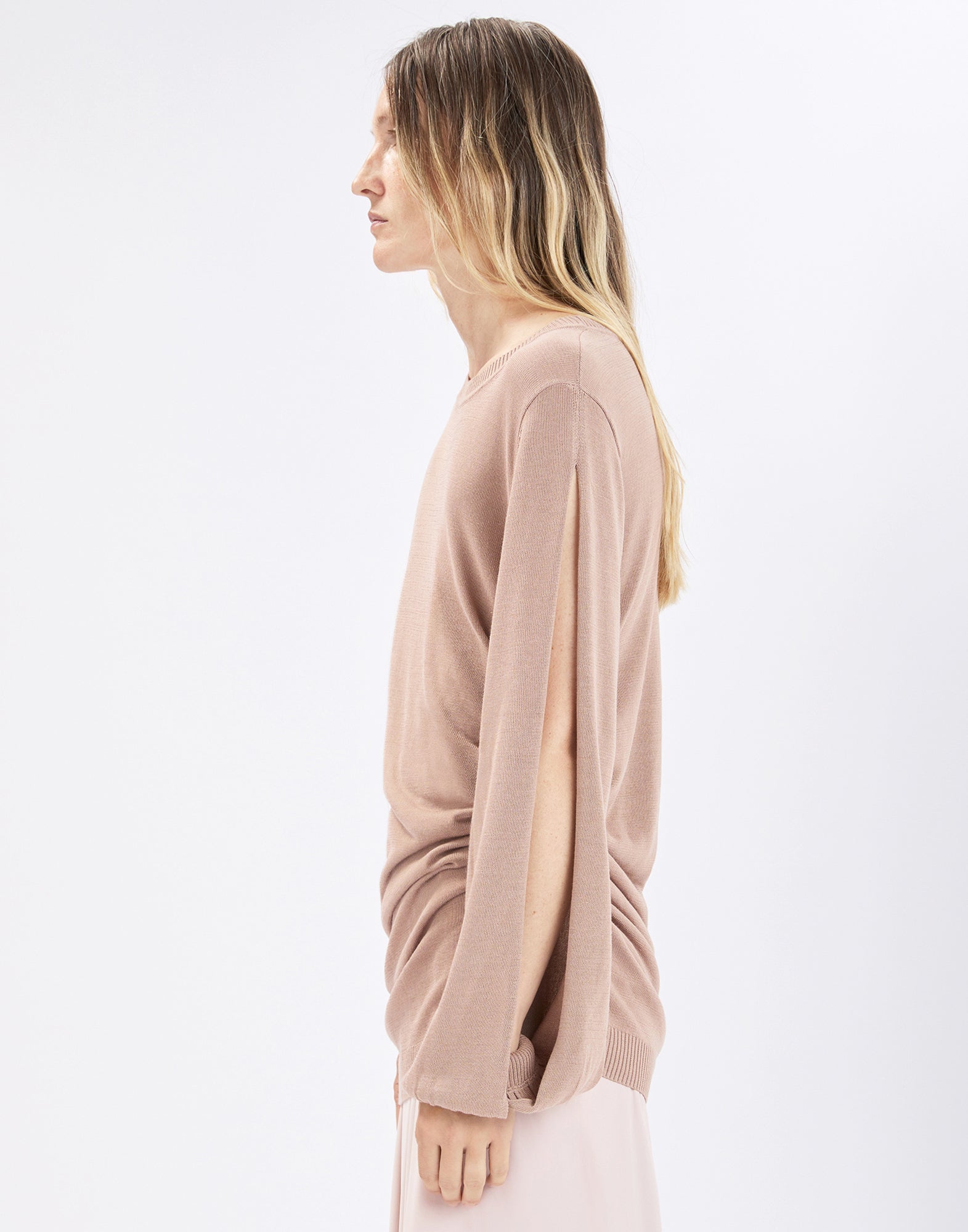 Side view: model wearing the sand-colored SANDUST knit sweater with crew neckline, loosely draped long sleeves, and a full-length opening along the outer side of the arm over a light-colored skirt.