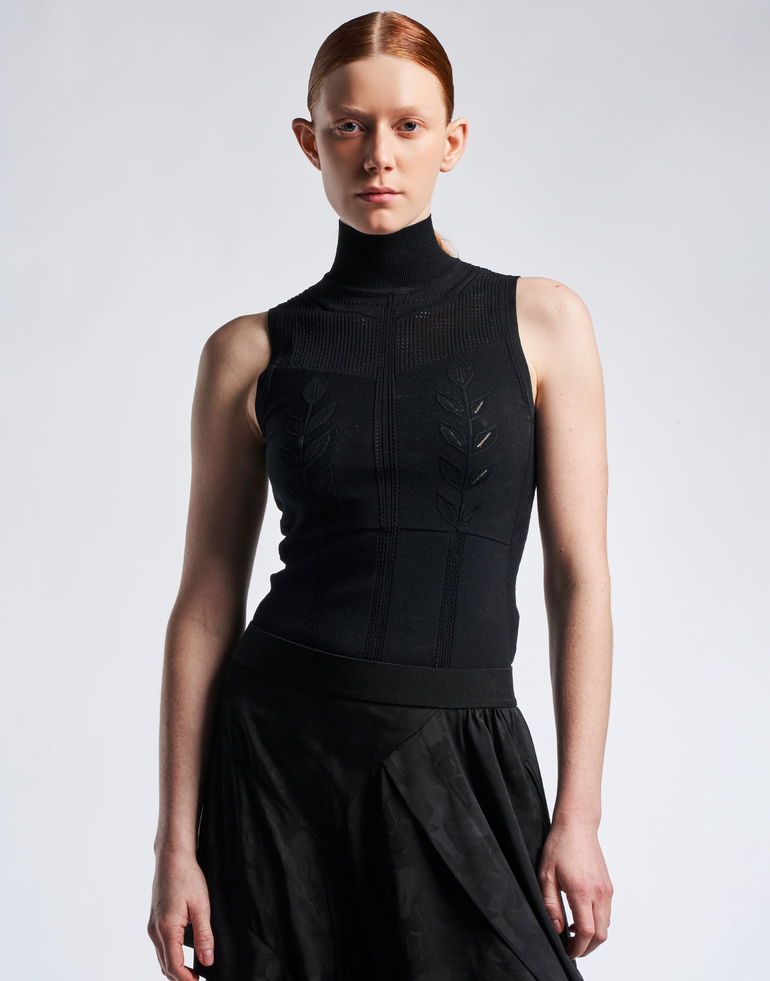 Front view of a woman wearing the sleeveless black GUISE top with high collar. Fine ribbed collar, perforated floral pattern on front, stretchy knit material.