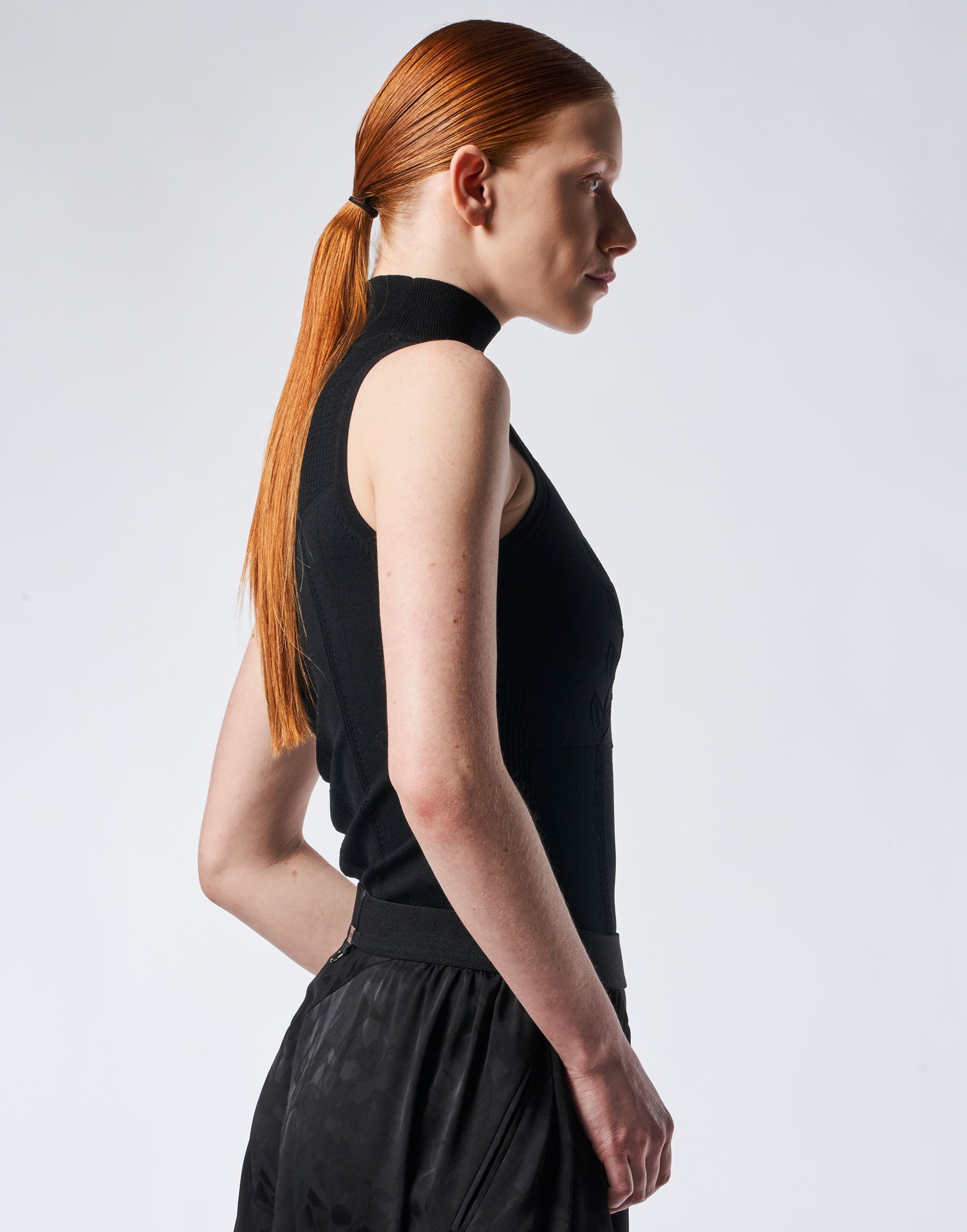 Side view of GUISE: model wearing sleeveless top with high fine ribbed collar in black, technical knit and perforated floral pattern.